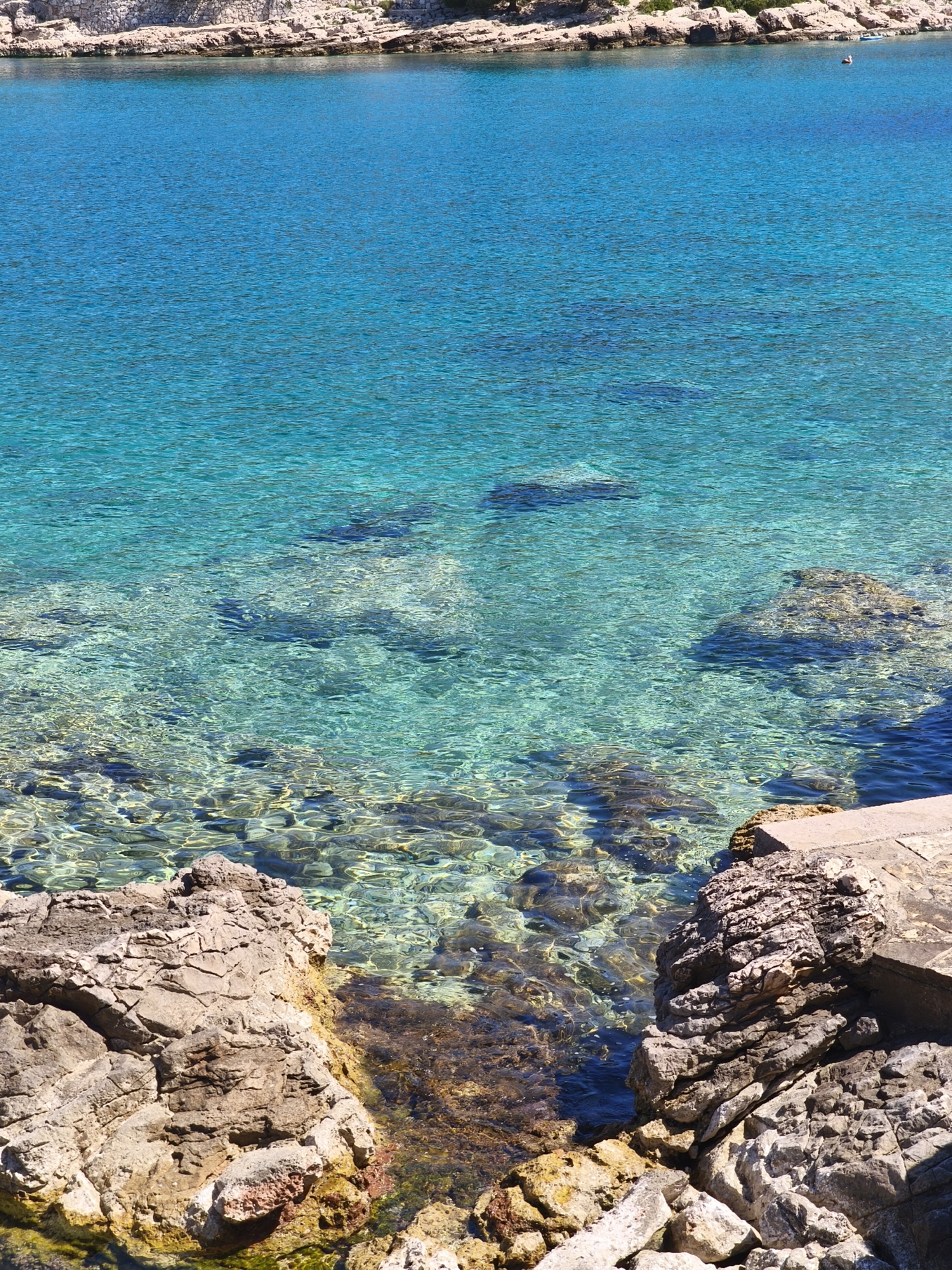 The image captures a clear, turquoise body of water meeting a rocky shoreline. The water transitions from a deep blue further out to a lighter, more transparent shade near the shore, revealing rocks and pebbles beneath the surface. The shoreline consists of jagged, tan-colored rocks and a concrete structure on the right. The location is Unknown, Unknown. There are no people visible in the immediate foreground. The scene is static, with no apparent action taking place.