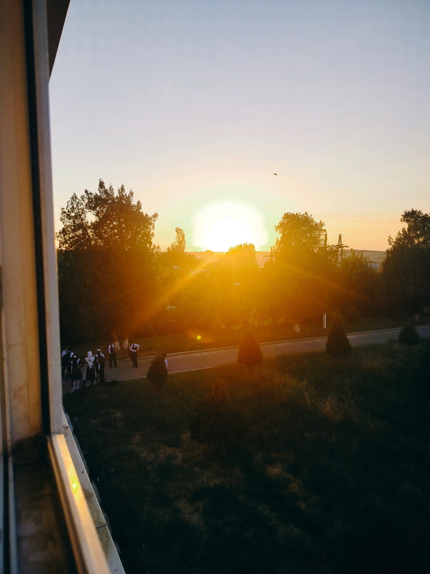 A group of young people in school uniforms are gathered on a paved area next to a grassy field, with a road visible in the background. The setting sun casts a warm, golden light across the scene, with trees silhouetted against the bright sky. The sun's rays create lens flares and a vibrant glow around the horizon. The image appears to be taken from a higher vantage point, looking out from a window or balcony. The overall atmosphere is serene and captures the end of a day. The location is identified as the city of Kölkent, Kazakhstan.