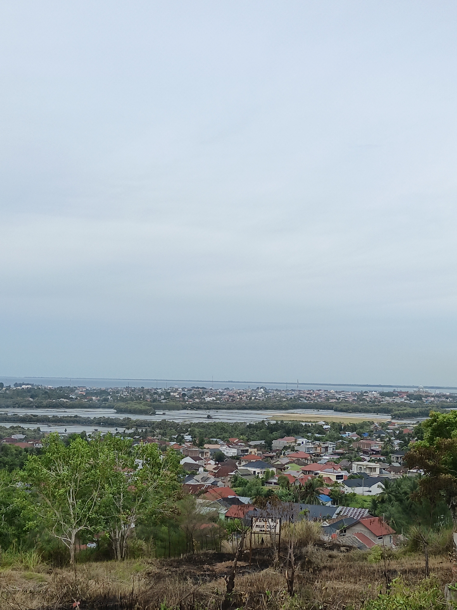 The image depicts an elevated view of a coastal urban area in Lhokseumawe, Indonesia, on an overcast day. In the foreground, a patch of dry, partially burnt vegetation slopes down towards a cluster of residential buildings with diverse roof colors, predominantly red and brown. Further back, a series of shallow, possibly agricultural, water bodies are segmented by strips of land and vegetation, leading towards a thin strip of coastline. Beyond the coastline lies a densely populated area extending along the shore. The overcast sky takes up the majority of the image. No people are visible.