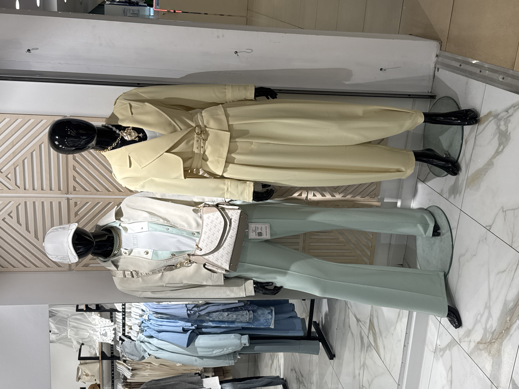 Two mannequins are displayed in a shop window, likely in Saint Petersburg, Russia, based on the style and potential location of similar businesses. The upper mannequin wears a light yellow belted coat-dress with a gold necklace. The lower mannequin is dressed in a white jacket, a white and pale blue shirt, light blue pants, and a white baseball cap. A patterned white and gray handbag hangs off the lower mannequin's shoulder. A clothing rack with clothes in similar colors is visible in the background. The floor is white marble.