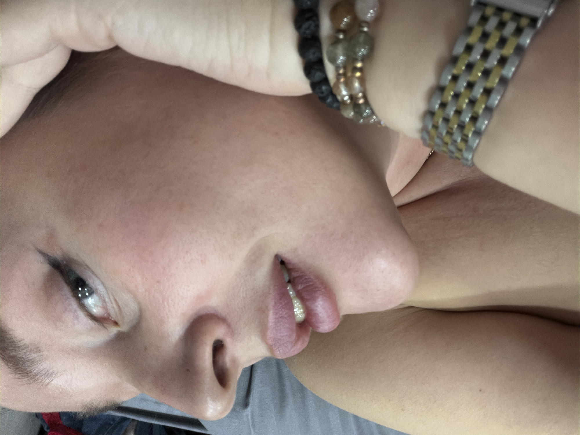 A close-up, angled view of a person's face, neck and arm wearing a watch and beaded bracelets. The person has light skin, defined eyebrows, eye liner, and slightly parted lips, revealing the upper teeth. The background is blurry but appears to be a gray sheet. The location is not discernible from the image.