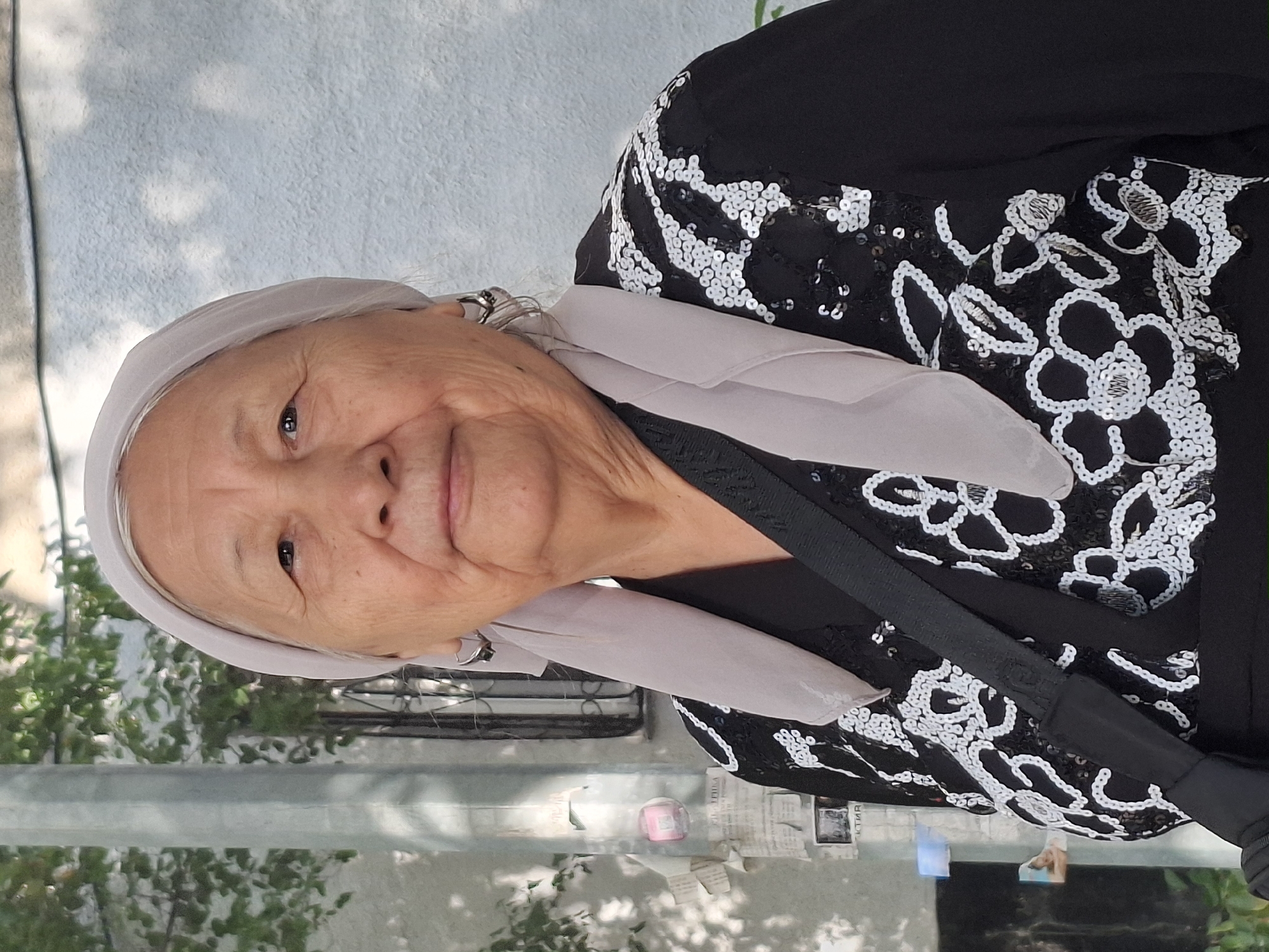 An elderly woman wearing a light purple headscarf and a black top with white floral embroidery and sequins is pictured in a close-up portrait. She has hoop earrings and a dark strap across her chest. The background consists of a light-colored textured wall and some greenery, suggesting an outdoor urban setting. There are also some posters on a pole to the left of the woman. The lighting indicates it is daytime. The scene is likely located in the city of Almaty, Kazakhstan, due to the presence of Cyrillic script on the posters.