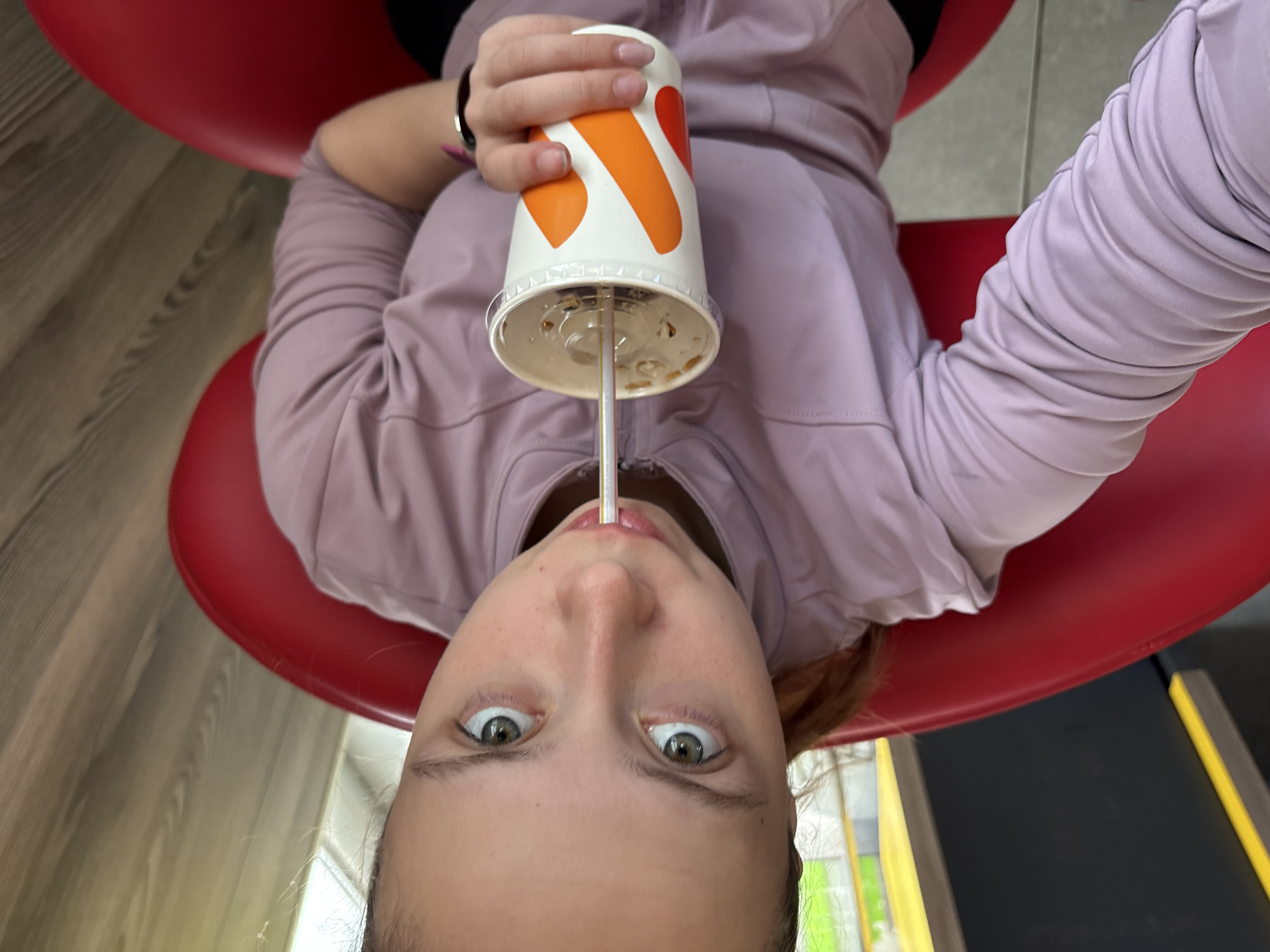 A woman is seated in a red chair, seen from a low angle. She is wearing a light purple long-sleeved shirt and a black watch. She is holding a large white paper cup with orange and white designs. A straw is in her mouth, suggesting she is drinking. The location is Perm, Russia, based on the cup's design. A wooden floor is visible in the background, along with elements of a wall with a yellow stripe.