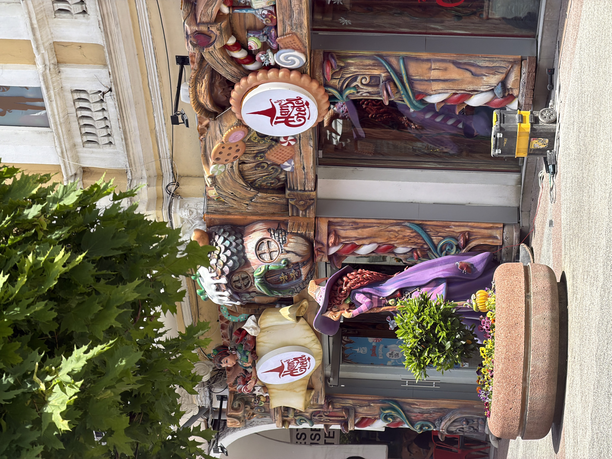 The image shows the facade of a building in Plovdiv, Bulgaria, possibly a confectionery or sweet shop, given its whimsical decoration. The exterior is adorned with elaborate, cartoonish carvings of gingerbread figures, candies, and other fairytale elements. A sign with the inscription "Hansel & Gretel" in red letters is visible. A planter with greenery and flowers sits in front of the entrance. Part of a tree is seen on the left side of the image, obscuring a portion of the facade.