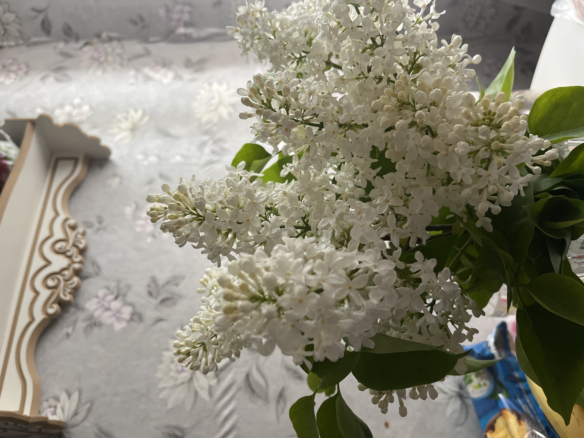 A close-up shot reveals a bouquet of white lilacs with green leaves, partially obscuring a floral-patterned surface. To the left, a decorative shelf with ornate carvings is visible. The location is identified as Мшинская, Russia.