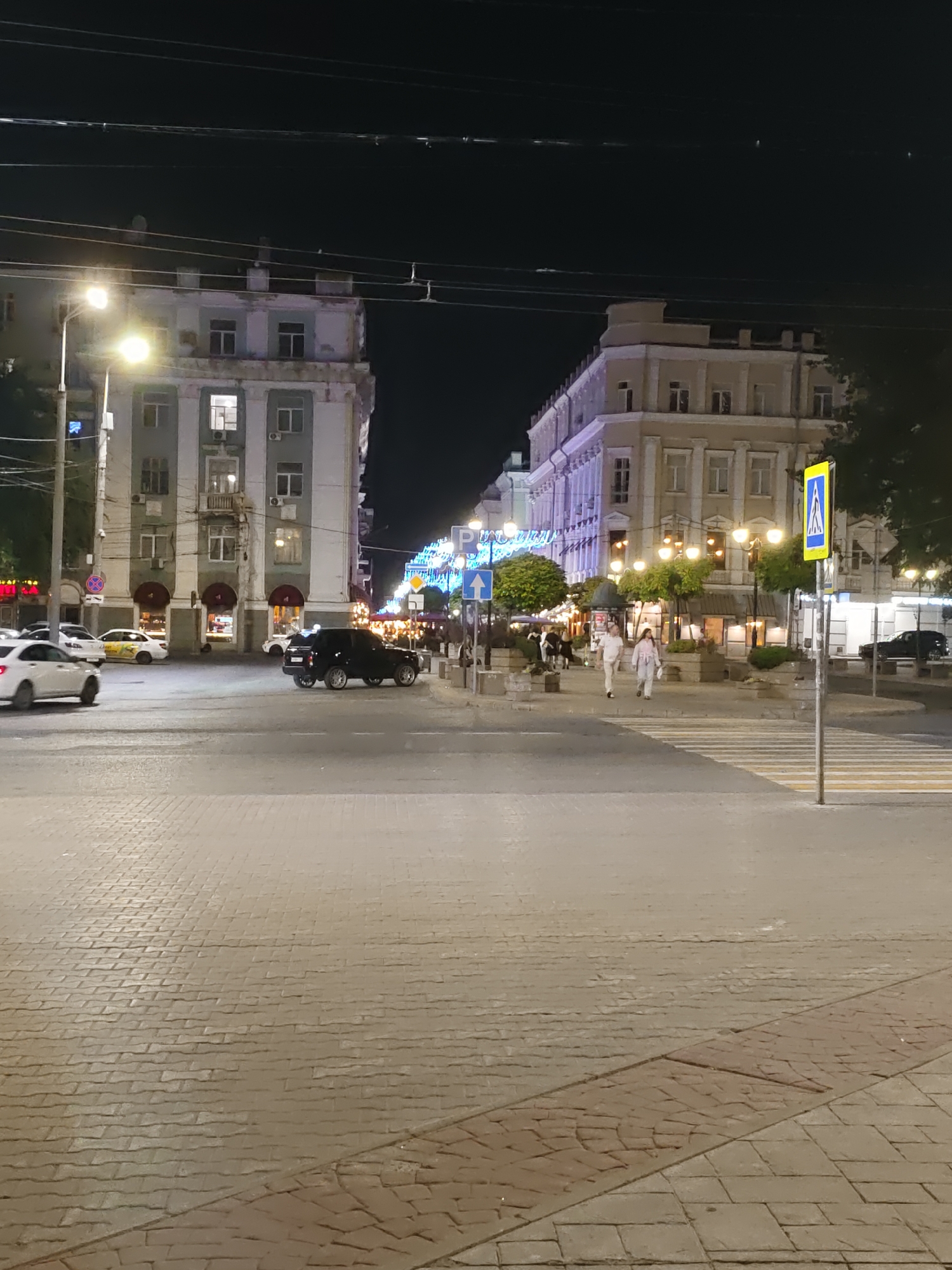 The image depicts a nighttime street scene in Rostov-on-Don, Russia, likely in the city center. A wide, paved intersection is visible, with cobblestone pavement in the foreground and a pedestrian crossing marked with yellow and white stripes. Two buildings flank the street. The building on the left appears to be older, with a light blue and white facade, multiple windows, and what seem to be shops or cafes at street level with arched entrances. The building on the right is a lighter color, possibly cream or beige, with a more ornate facade featuring columns and multiple windows.
Several vehicles are present: a white car and a yellow car are parked or moving on the left side of the intersection. A black SUV is positioned in the middle of the intersection. In the background, more cars are visible.
Two people, a man and a woman, are walking across the pedestrian crossing on the right side of the image. They are dressed in casual, light-colored clothing.
Streetlights illuminate the scene, casting a warm glow. Decorative lights, possibly string lights or illuminated signs, are visible on the building facades in the distance, adding to the nighttime ambiance. Trees and planters are also integrated into the streetscape. Power lines are strung across the top of the image. A yellow and blue pedestrian crossing sign is posted on the right side of the street. The overall impression is of a lively urban environment at night.