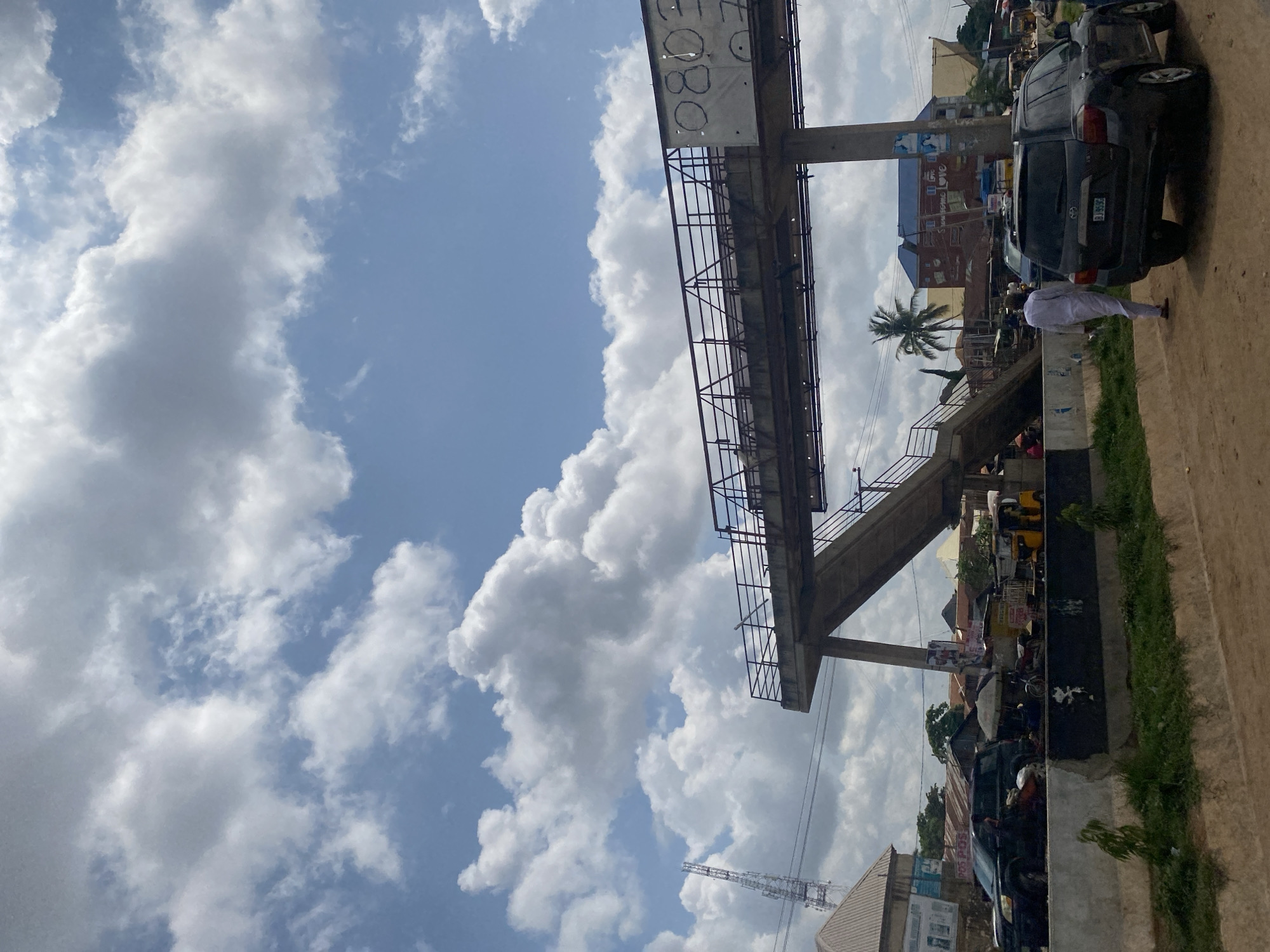 In Ado, Nigeria, an overpass under construction spans a busy street. The concrete structure features a walkway with railings, supported by a large pillar and an angled ramp. In the foreground, a dirt path runs alongside a grassy verge. A dark SUV is parked on the side of the road, and a person in white attire walks away from it. Further down the street, a yellow tricycle, a motorcycle, and various market stalls are visible. Buildings with corrugated iron roofs and brightly colored facades line the street. A tall telecommunications tower stands in the background, silhouetted against a sky filled with dramatic, cumulus clouds.