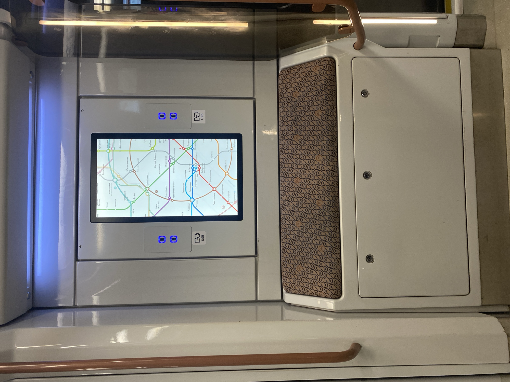 An interior view shows a section of a public transport vehicle, likely a train, in an unknown location in Unknown, Unknown. The central focus is a digital display screen showing a subway map. Below the screen, there is a padded seating area with a repeating pattern of stylized "M" logos. Beneath the seating is a plain, off-white panel with two circular latches. To the left of the screen, there are two illuminated blue buttons. A USB symbol is also visible below the screen, indicating charging ports. A copper-colored grab bar is positioned to the left, and another to the right is partially visible. The overall environment appears clean and modern, consistent with an indoor transit setting.