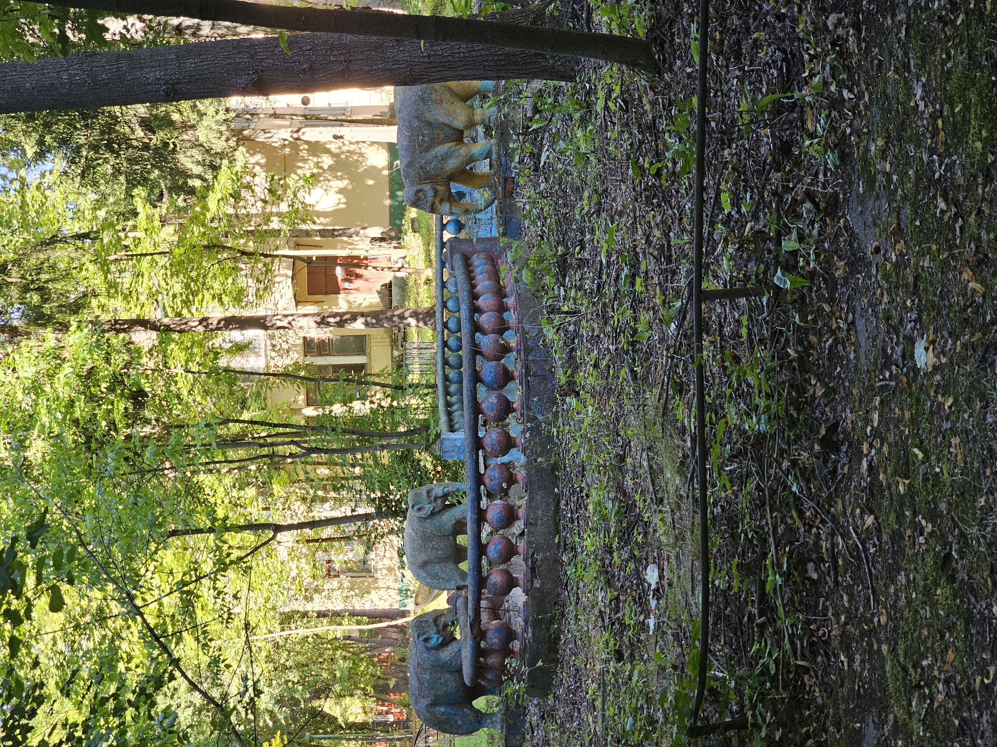 The image captures a weathered, outdoor scene with three elephant statues positioned behind a decorative, balustrade-like barrier. The statues appear aged and somewhat worn, with faded paint in shades of gray, blue, and yellow. The balustrade itself is also distressed, featuring a pattern of spherical elements painted in red and blue. The surrounding environment is overgrown and appears somewhat neglected. Dense foliage, including trees with lush green leaves, dominates the background, with a building visible behind the trees. The ground is covered in leaf litter, twigs, and patches of green vegetation. The overall impression is one of an abandoned or forgotten space, possibly a park or playground, with elements of decay and nature reclaiming the area. The location is identified as Khimki, Russia.