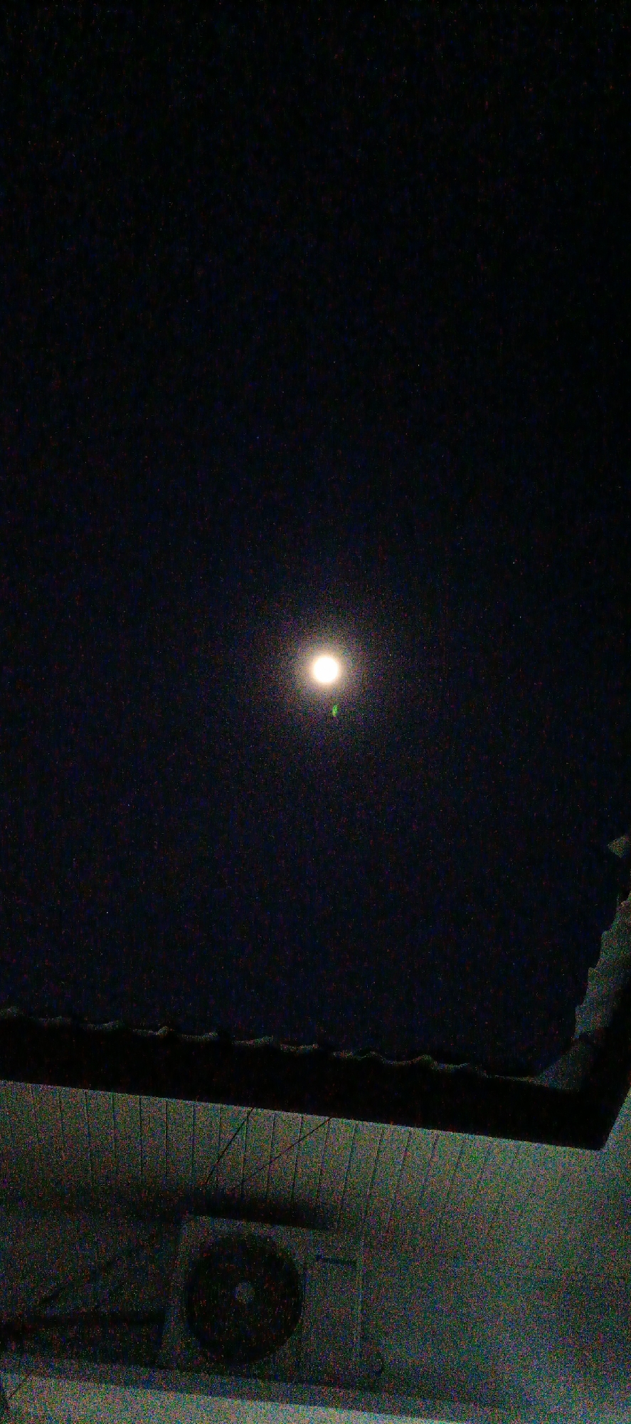 The image depicts a nighttime scene from a low-angle perspective, looking upwards. The dominant feature in the sky is a bright, round light source, possibly the moon, emitting a strong glow. This light is surrounded by numerous small, faint specks, which are likely stars. Below the sky, a dark, shadowed structure, possibly the eaves of a building or roofline, is visible. Further down, an air conditioning unit is mounted on a wall, indicated by a rectangular casing with a circular fan grille. The overall image is grainy, suggesting a low-light photograph. There are no people or identifiable landmarks of Pashiebeutong, Indonesia present in the image.