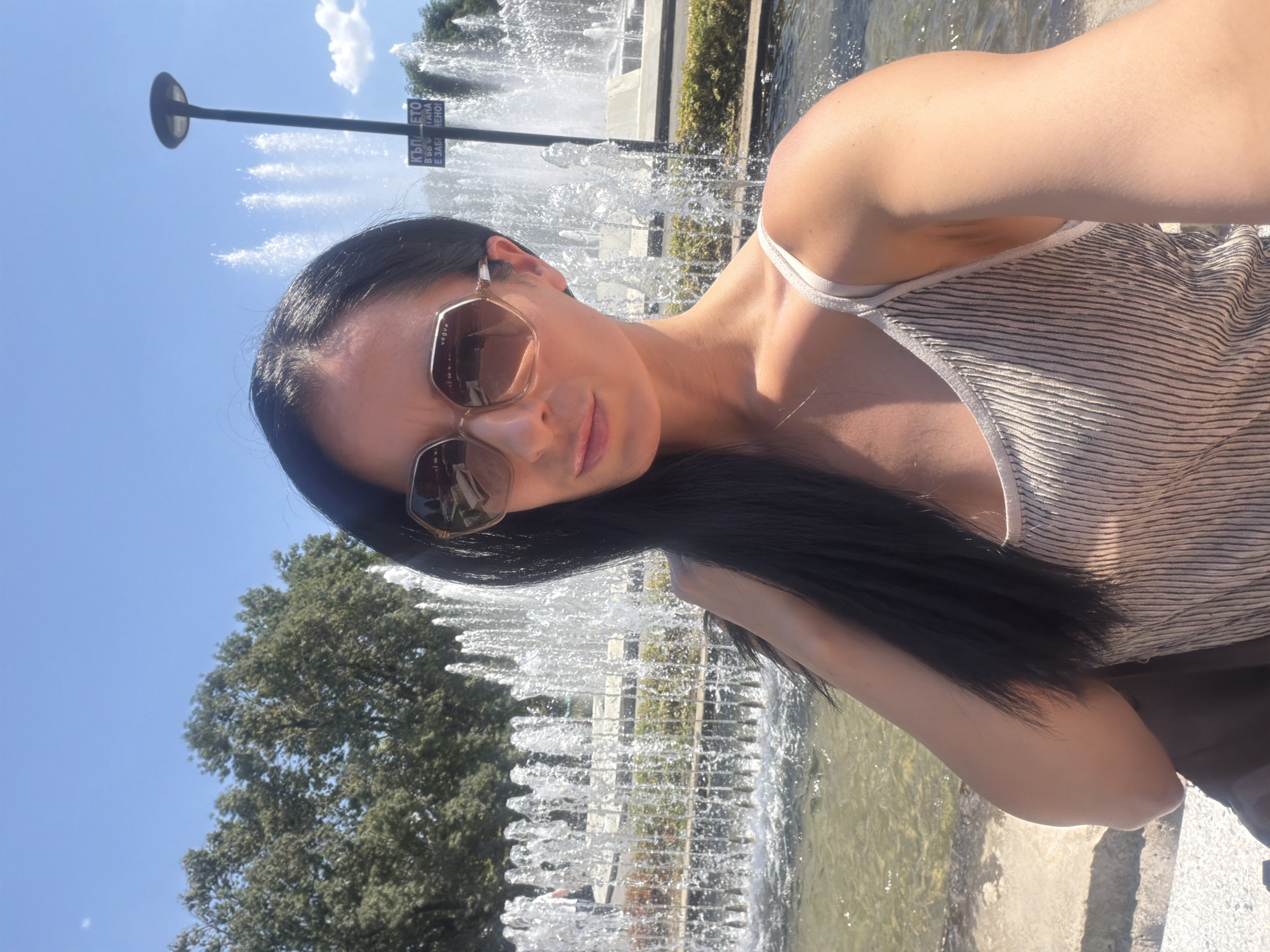A selfie is taken by a woman with long, dark hair and sunglasses in front of a fountain in Pleven, Bulgaria. The woman is wearing a striped tank top. The fountain is spraying water high into the air, and there are trees in the background. A street lamp and a sign with Cyrillic writing are also visible. The sky is clear and blue, and the sun is shining.
