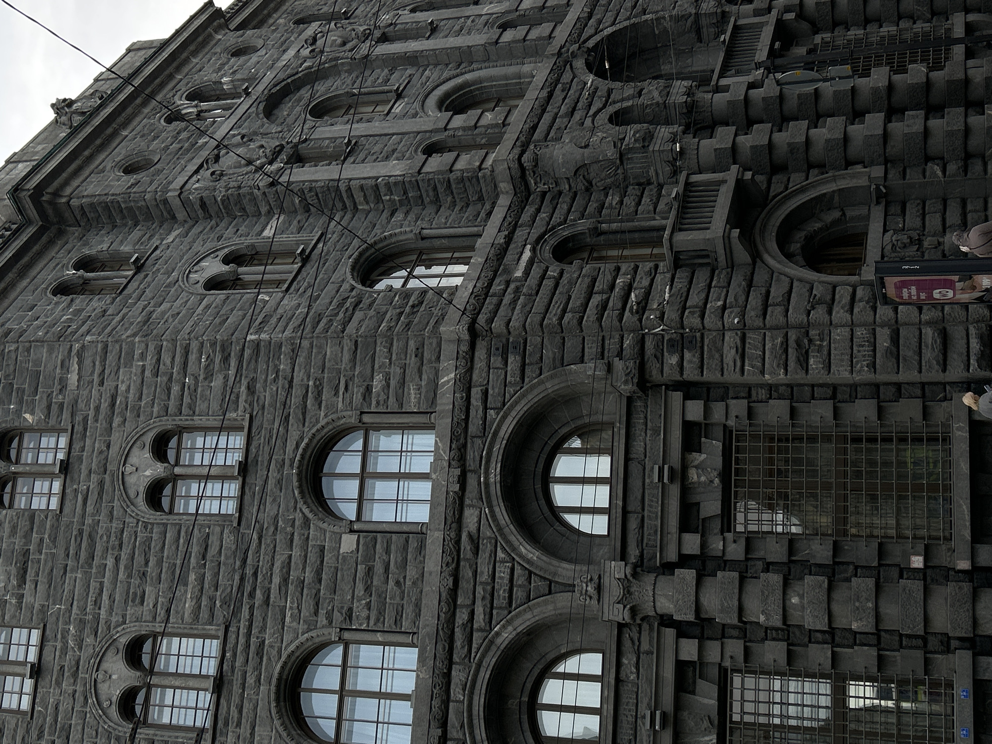 The image shows a facade of a large, dark grey stone building in Saint Petersburg, Russia. The architectural style appears to be historic and ornate, with multiple rows of arched windows. The windows on the left side of the facade are rectangular. The windows on the right side of the facade are round. The stonework is textured, with a rough-hewn, block-like appearance. Some areas of the facade are more detailed, with decorative elements built into the stone. The right side of the facade includes what appear to be external stairs or balconies integrated into the building's design, along with a large cage-like structure covering a window. A small part of a person's head can be seen walking on the street level on the right side of the image. Utility wires are visible against the sky, running diagonally across the upper portion of the facade. The overall lighting suggests overcast or cloudy weather.