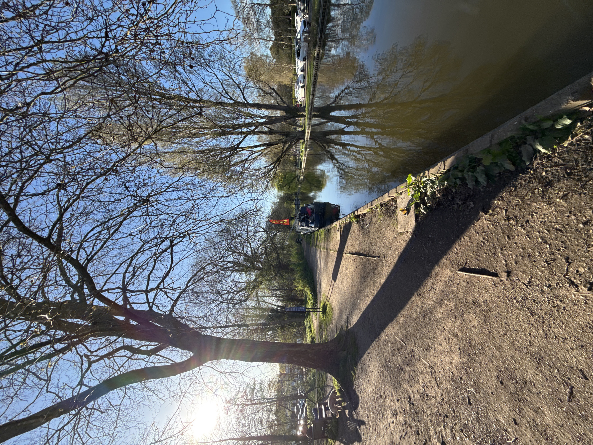 The image shows a canal-side scene in Dacorum, United Kingdom. The canal is bordered by a concrete wall on one side and a dirt path on the other. Trees with bare branches line both sides of the canal, and their reflections are visible in the water. A narrowboat is moored along the canal, with an orange flag visible. On the path side, there's a prominent tree trunk casting a long shadow. The path appears to be made of compacted dirt or gravel. In the background, parked cars can be seen along the canal. The sky is clear and blue.