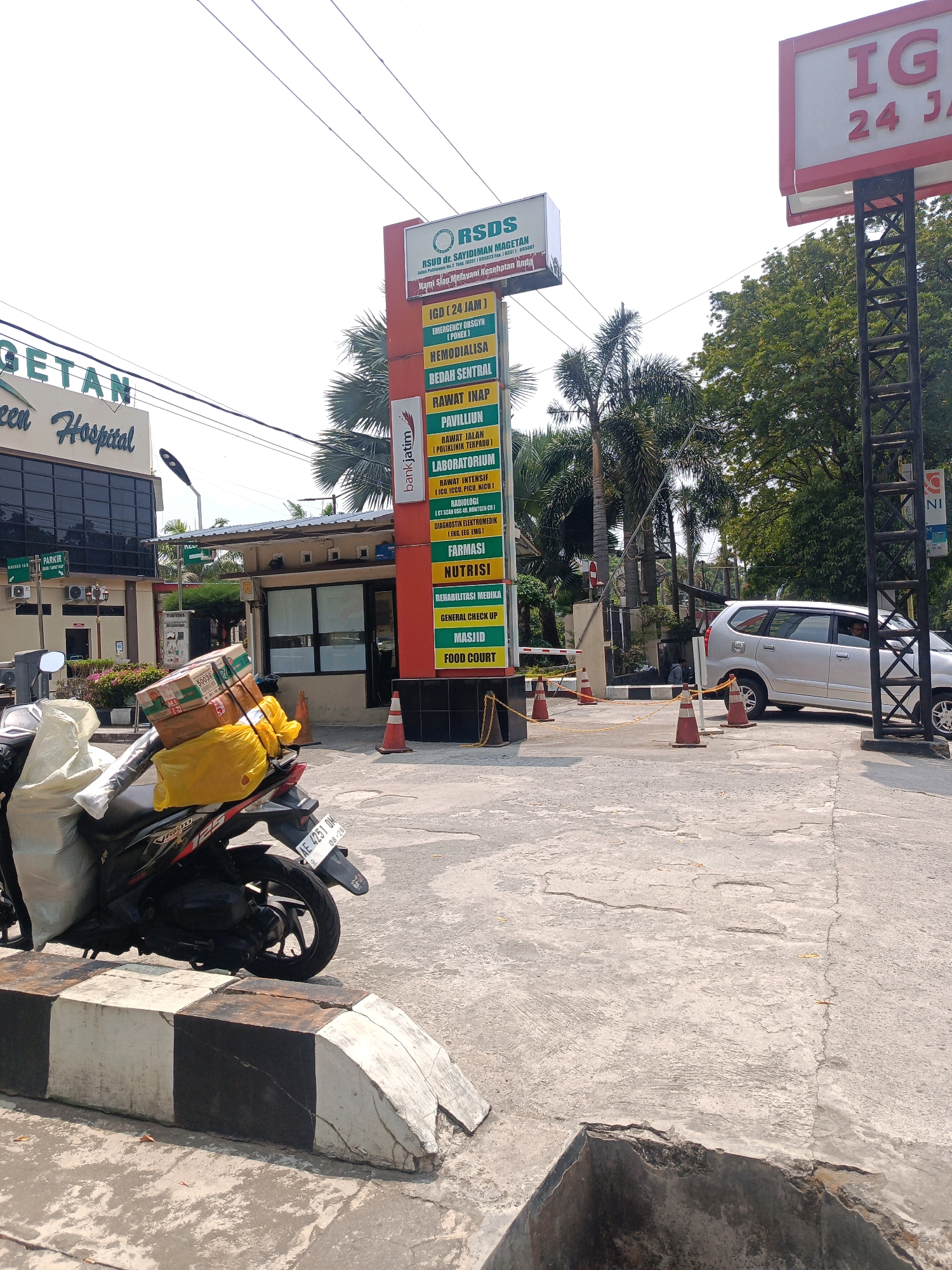 The image shows the entrance to the RSUD Dr. Sayidiman Magetan hospital in Tambran, Indonesia. A vertical sign lists various hospital services, including IGD (24 JAM), EMERGENCY OBSGYN, HEMODIALISA, BEDAH SENTRAL, RAWAT INAP, PAVILLION, RAWAT JALAN (Poliklinik Terpadu), LABORATORIUM, RAWAT INTENSIF (ICU, ICCU, NICU), RADIOLOGI (CT Scan, USG, Rontgen), DIAGNOSTIK ELEKTRODIAGNOSTIK (EKG, EEG, EMG), FARMASI, NUTRISI, REHABILITASI MEDIKA, GENERAL CHECK UP, MASJID, and FOOD COURT. A motorcycle loaded with boxes and a yellow bag is parked in the foreground. A silver minivan is entering the hospital grounds through a gate controlled by traffic cones and a chain. The background features trees and a bright sky. Another sign above the hospital entrance reads "GETAN Hospital."