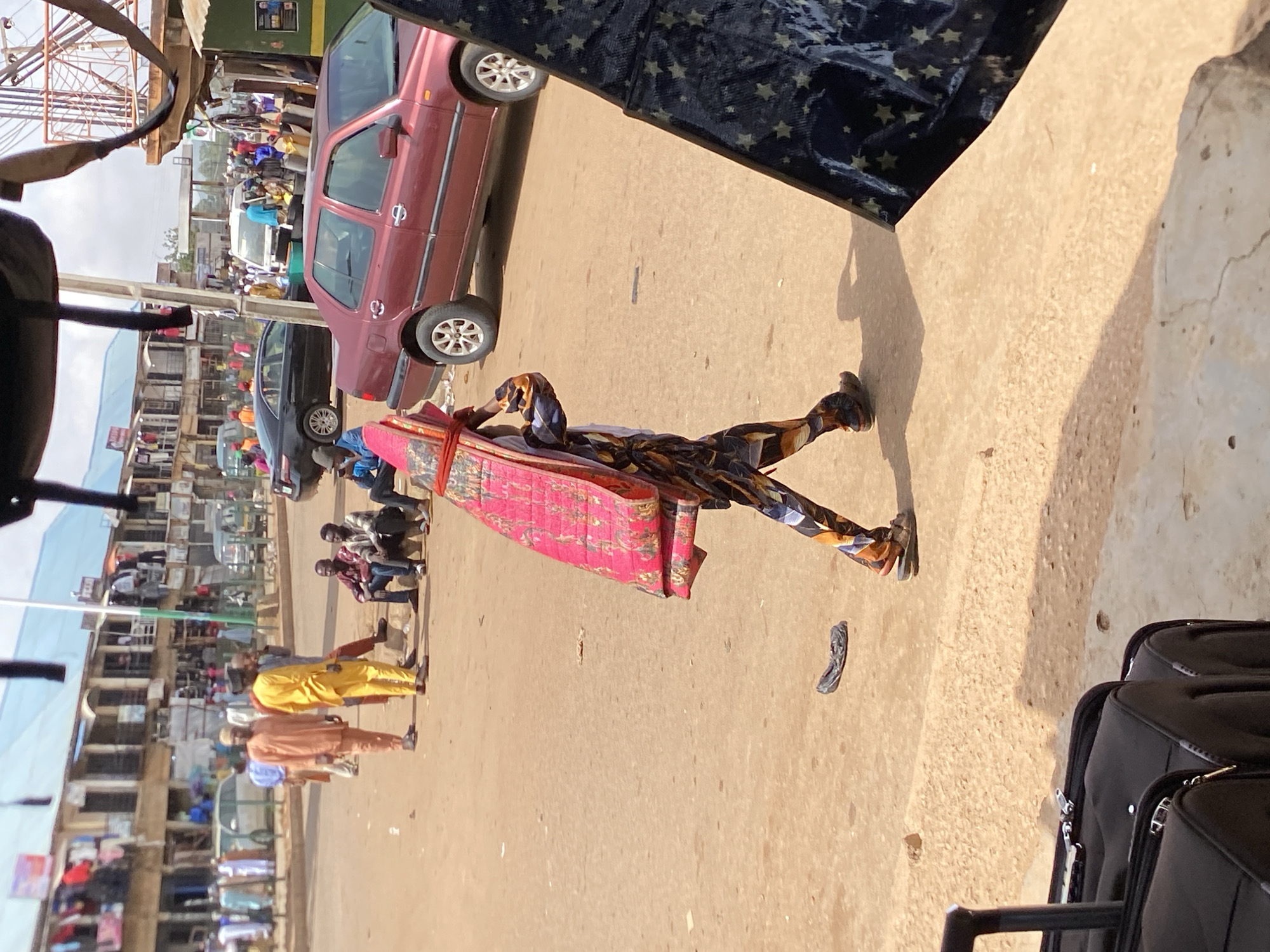 In Bukuru, Nigeria, a man wearing multicolored patterned clothing carries a red mattress on his head along a street. A maroon car is parked on the side of the road. Other people are visible in the background near buildings with multiple stories, and some are sitting on benches. Suitcases sit on the ground in the foreground. A sandal lies on the ground in front of the man.