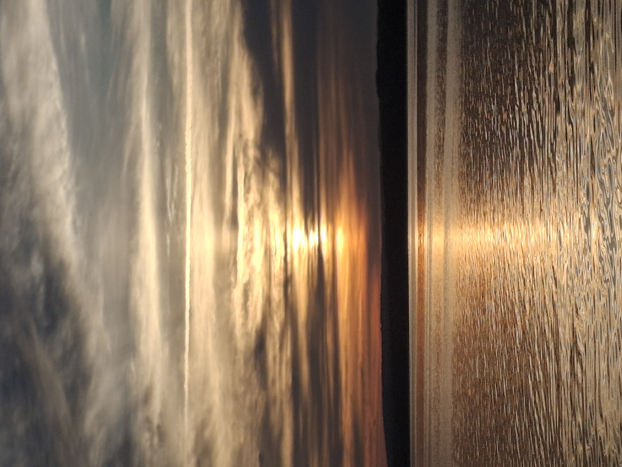 The image depicts a serene sunset over a body of water, likely the Kama River, in Chaikovsky, Russia. The sky is filled with horizontal bands of clouds, transitioning from dark grays and blues at the top to warm oranges and yellows closer to the horizon, where the sun is partially obscured. The sun's rays create a shimmering golden path across the rippling water. A dark, silhouetted landmass, possibly the riverbank or distant hills, forms a distinct line separating the sky from the water. The overall scene is one of tranquil natural beauty, characteristic of Chaikovsky's landscape.