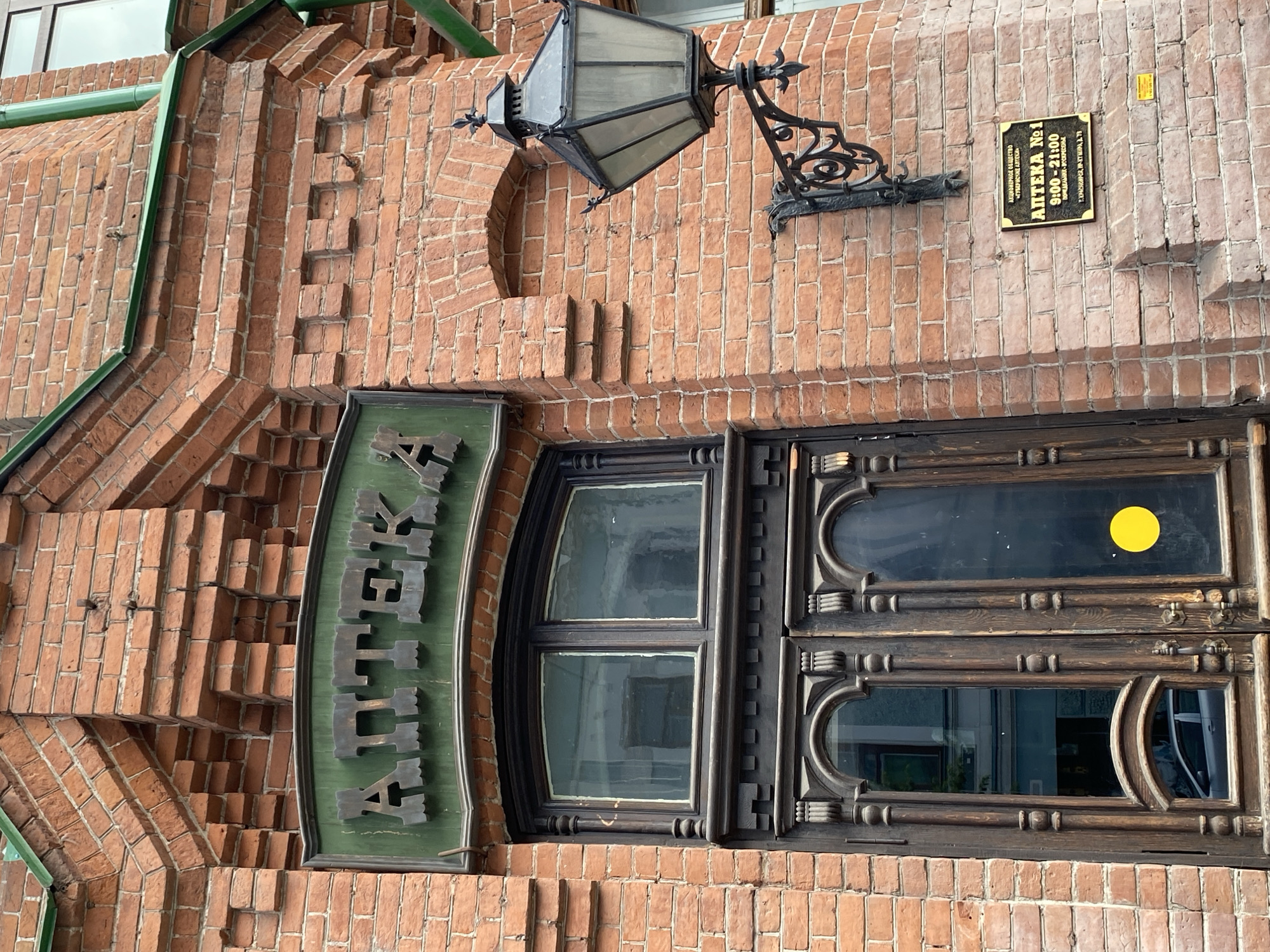 The image shows the facade of a brick building with an ornate wooden door and window. Above the entrance, a green sign with stylized lettering reads "АПТЕКА" (Apteka), which means "pharmacy" in Russian. To the right of the door, a wrought-iron lantern is mounted on the brick wall. Below the lantern, a rectangular gold sign displays information in Russian, including "АПТЕКА №1" (Apteka No. 1), operating hours "9:00 - 21:00," and an address "КРАСНОЯРСК, ПР. МИРА, Д. 71" (Krasnoyarsk, Mira Ave., Bldg. 71). This indicates the pharmacy is located in Krasnoyarsk, Russia, on Mira Avenue, building 71. The building's brickwork is intricate, with decorative elements and a green roofline visible at the top. The overall scene depicts the entrance to a historic pharmacy in a city.