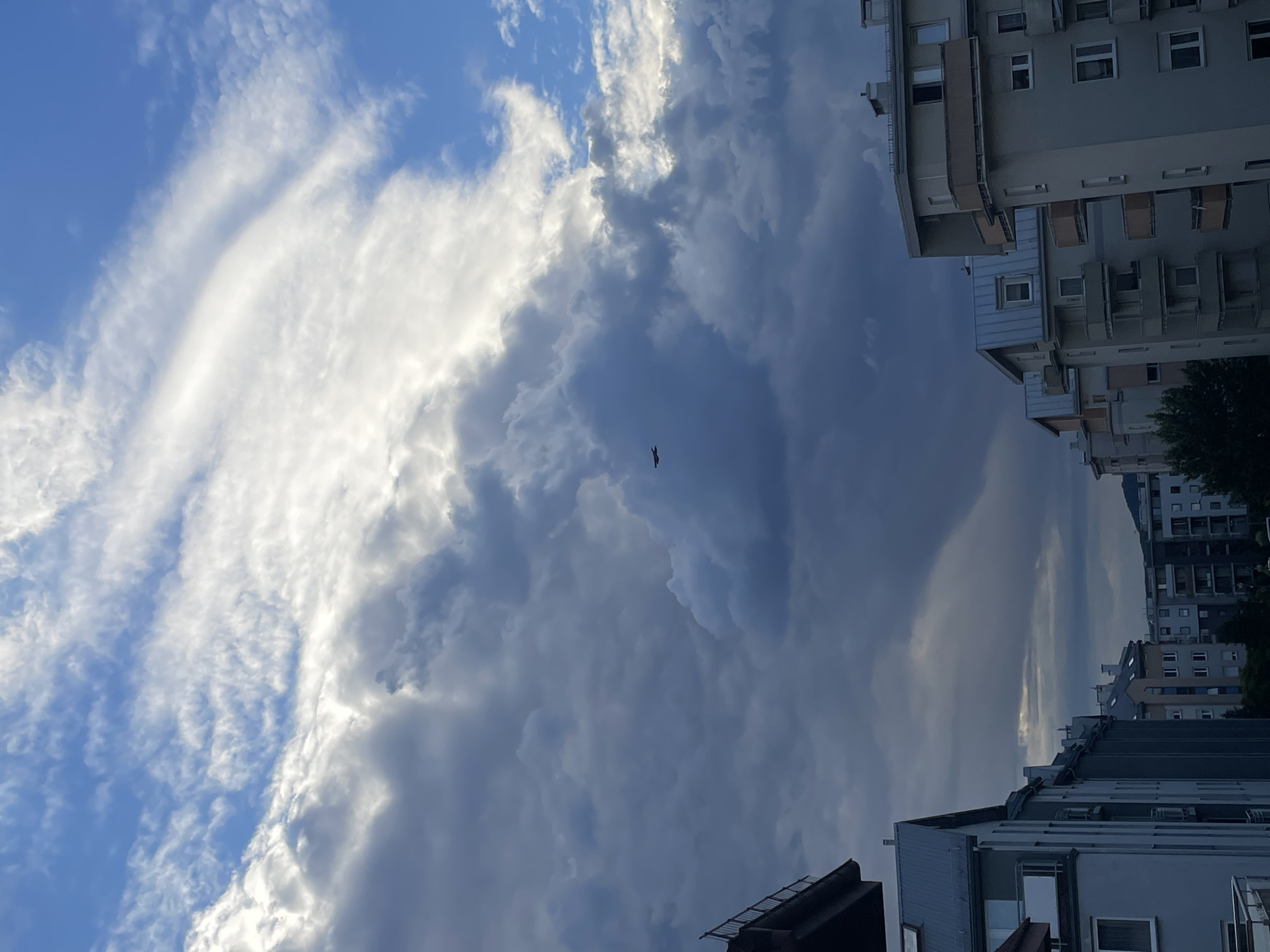 A single, small, dark-colored airplane is visible flying through a cloudy sky above a city. The sky is a mix of bright white and dark gray clouds, with patches of blue sky visible. In the foreground, the tops of several apartment buildings are visible. The buildings are modern in design, with multiple stories and balconies. One building on the left has a metal railing on a balcony. The overall impression is of a cloudy day in a residential area of a city.