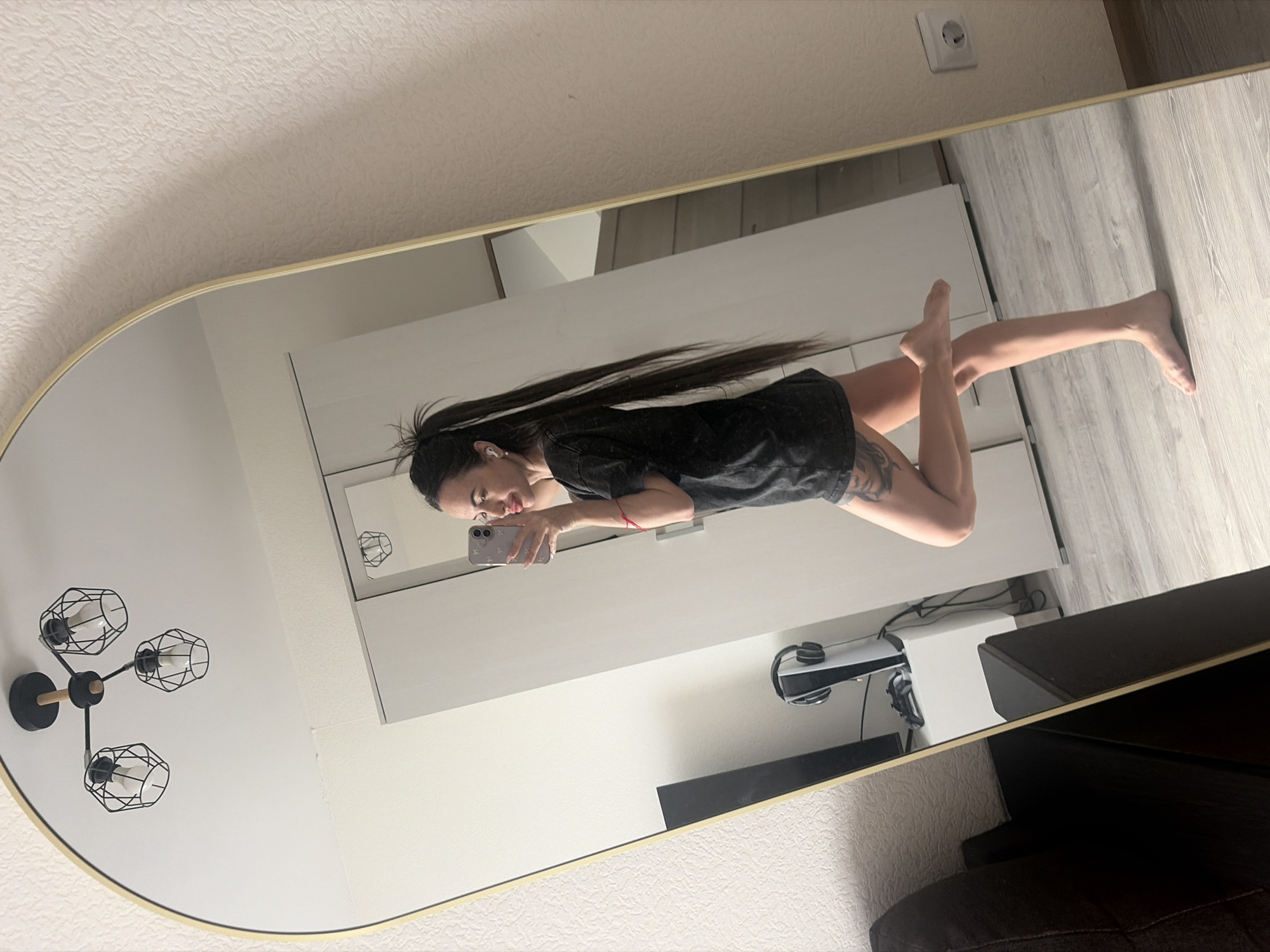 A medium shot depicts a woman reflected in an oval mirror. The woman is holding a smartphone and appears to be taking a photo of herself. She has long dark hair, fair skin, and tattoos visible on her arm. She is wearing a dark shirt. Her legs are visible, extending towards the right side of the frame.
The reflection shows a modern interior. A light fixture with a black and gold finish is on the left wall. A white dresser is in the background, with what appears to be electronic equipment, possibly a PlayStation, and a dark-colored piece of furniture beside it. The flooring is a light gray.
The setting is likely inside an apartment. Based solely on visual data, there is no information related to the city of Saint Petersburg, Russia.