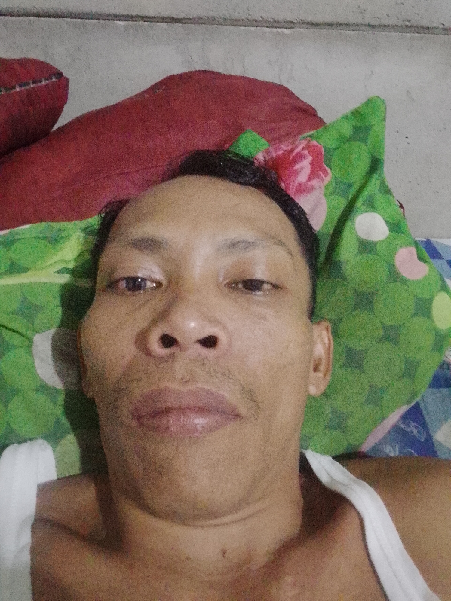 A selfie image of a man with dark hair, light brown skin, and dark eyes. He is lying on his back with a green, polka-dot pillow and a red pillow behind him. He is wearing a white tank top. The background appears to be a gray wall. I cannot determine any information about the location being Bukit Ketok, Indonesia, from the image itself.