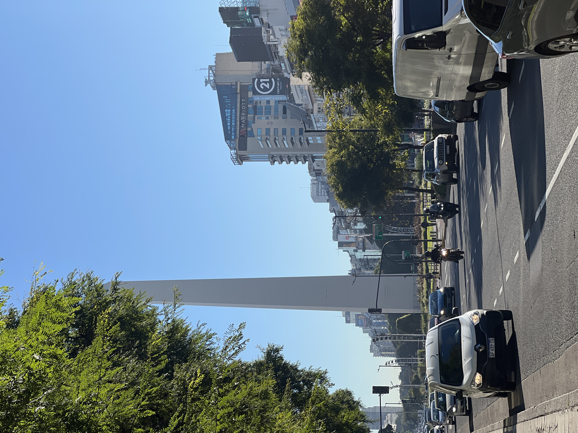 The image captures a street scene in Buenos Aires, Argentina, featuring the Obelisco as a prominent landmark in the background. Traffic is moving on the avenue, including cars and motorcycles. Tall buildings are visible along the avenue, including a Marriott hotel building with a "21" logo. The foreground includes trees alongside the road. The sky is clear and blue.