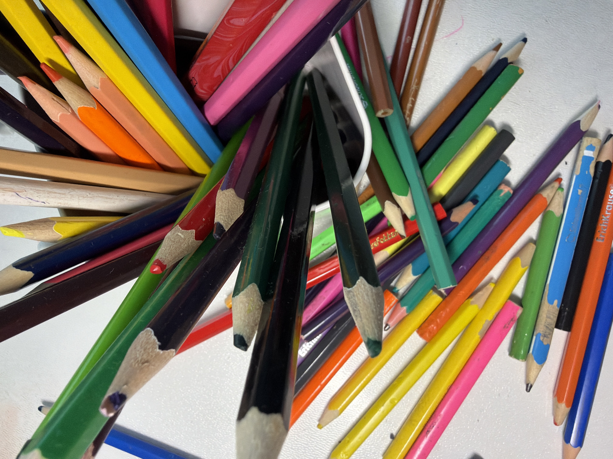 The image shows a collection of colored pencils of various lengths and colors. The pencils are scattered on a white surface. The color palette includes shades of green, blue, yellow, orange, brown, purple, pink, and red. Several pencils are partially sharpened. There is also an eraser visible among the pencils. The scene appears to be an indoor shot taken with natural lighting. There is no indication of the image being located in the city of Votkinsk, Russia. There are no people in the picture.