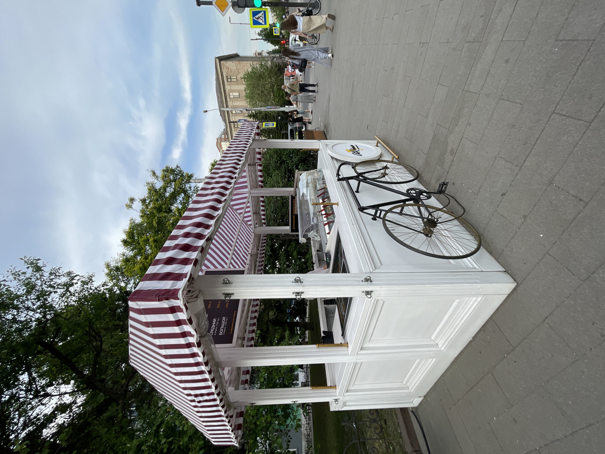The image depicts an outdoor scene in Krasnoyarsk, Russia, likely a city street or park. In the foreground, a white kiosk with a red and white striped awning is set up. The kiosk appears to be selling ice cream or similar treats, with a glass display case showing various flavors. A vintage-style black bicycle is leaning against the kiosk.
Several people are visible in the background, walking on a paved walkway. The environment includes trees and buildings, suggesting an urban setting. Traffic lights and street signs are also present, indicating a public thoroughfare. The sky is partly cloudy. The overall impression is of a commercial area on a pleasant day.