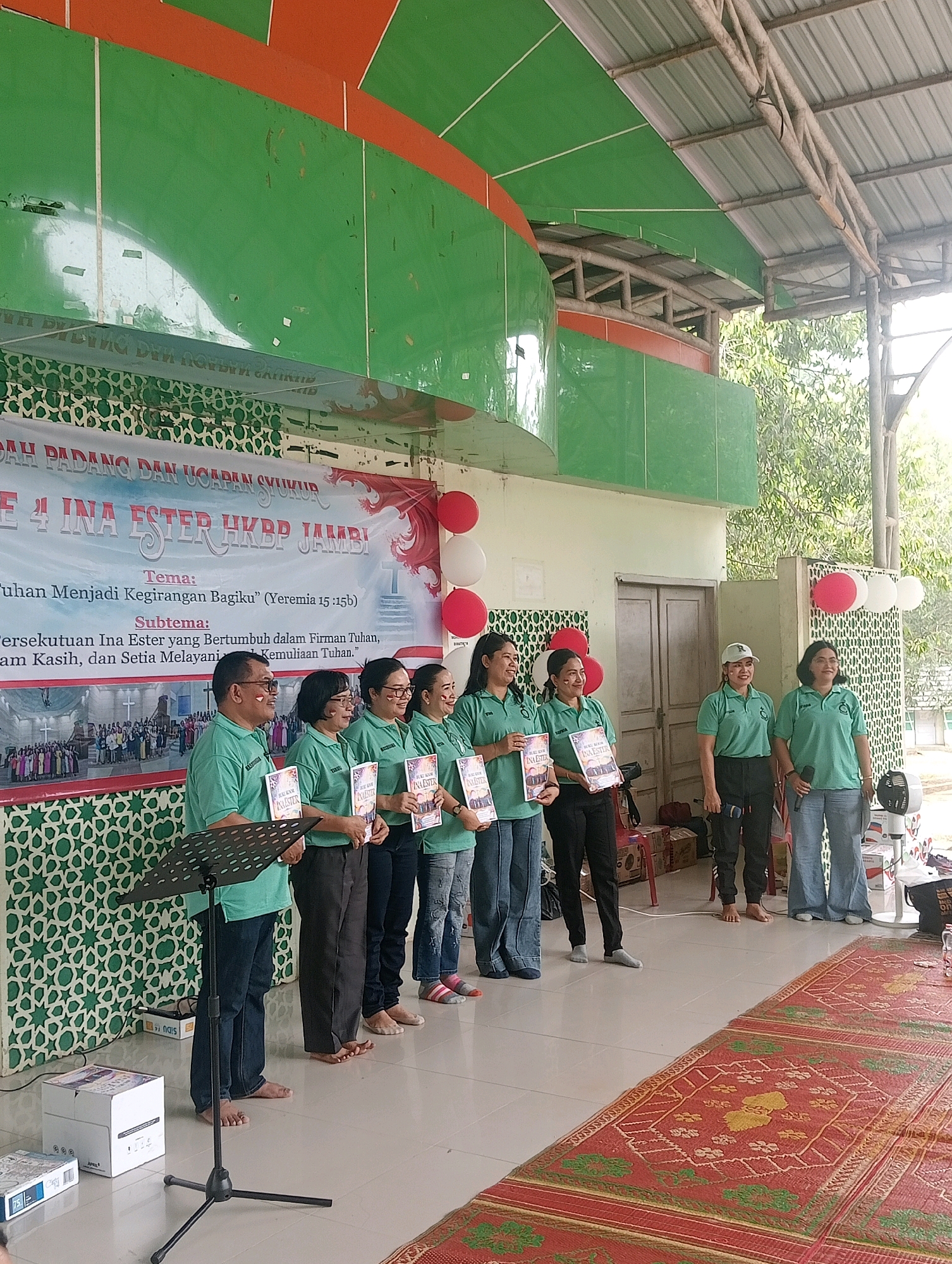 An outdoor stage area in Jambi, Indonesia, is set up for an event, likely a church gathering given the banner text. The stage has a green and orange curved roof structure overhead, supported by metal beams, with a corrugated metal roof. A white banner with red decorative elements is hung behind the group of people, displaying text in Indonesian, including "PUJIAN PADANG DAN UCAPAN SYUKUR" and "INA ESTER HKBP JAMBI." Below this, themes and sub-themes are listed.
A group of nine people, predominantly women, are standing on the stage, facing forward. They are all wearing matching light green polo shirts with a logo. Most are holding books or folders. Seven individuals are visible from the waist up, while two on the right are more fully visible. One man on the far left is also wearing a green polo shirt and dark pants. He is standing next to a music stand.
The backdrop behind the individuals is a white wall with a repeating green geometric pattern. Red and white balloons are attached to the right side of the banner. The floor is tiled with a large, colorful rug in the foreground. Several cardboard boxes are placed on the floor to the left of the stage.
The overall scene suggests a formal gathering, possibly a celebration or presentation, taking place in a public or semi-public outdoor space in Jambi.