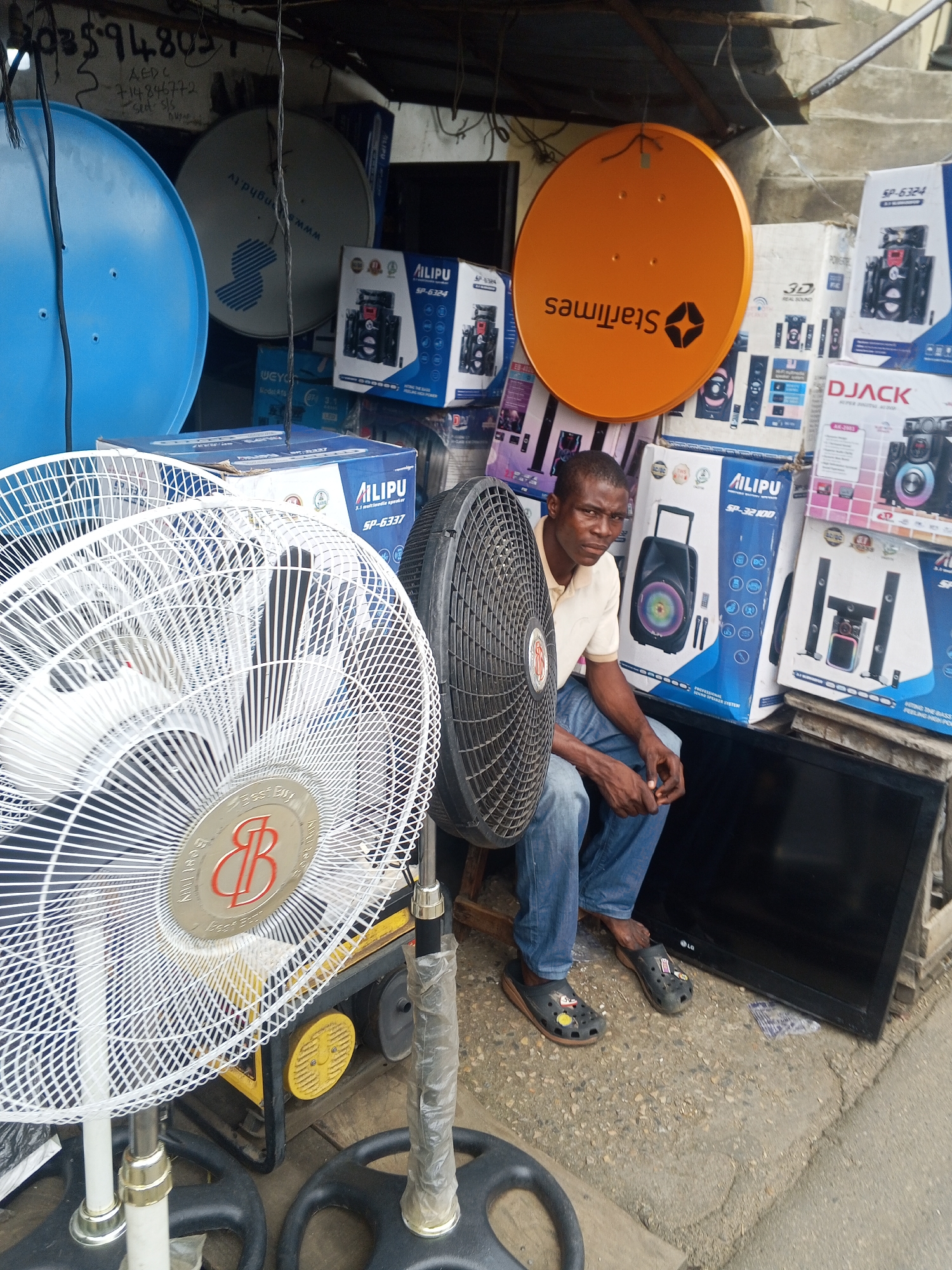 The image depicts a street vendor in Okene, Nigeria, surrounded by various electronic goods for sale. The vendor, a man wearing a light-colored polo shirt and blue jeans, sits on a stool next to a large black television. In front of him, two white oscillating fans are prominently displayed, with one partially obscuring a black fan behind him.
Behind the vendor, a stack of boxes containing audio equipment, likely speakers, are arranged. Brands such as "Ailipu" and "DJACK" are visible on these boxes, along with model numbers like "SP-6324" and "AR-2093". To the left, several satellite dishes are also present, including a bright orange one with the "StarTimes" logo and a silver one with a logo that appears to be "nghd.tv". A large blue satellite dish is also visible.
The setting appears to be an outdoor market or roadside stall, with the goods displayed under a makeshift shelter. The ground is a mixture of gravel and concrete. The overall scene suggests a commercial transaction is taking place or is about to take place, with the vendor awaiting customers.