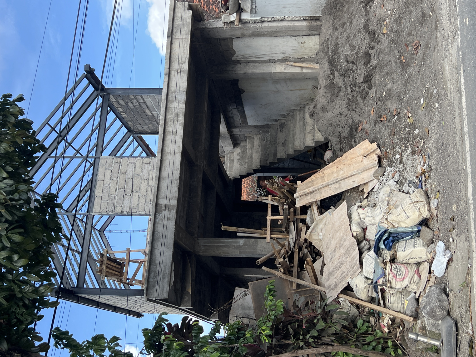 The image shows a construction site in what appears to be a tropical area, potentially within the Ubud District, Indonesia. The scene features the skeletal structure of a building under construction, with a steel frame visible against a clear blue sky. Concrete walls and levels are partially erected, with a staircase visible in the interior. A pile of construction debris, including wood planks, bamboo, and bags of cement, sits in the foreground, partially obscured by greenery. The ground is covered in dirt and debris. No people are immediately visible in the shot. Power lines run across the upper portion of the frame. The overall impression is one of an active construction zone within a lush, possibly urban, environment.