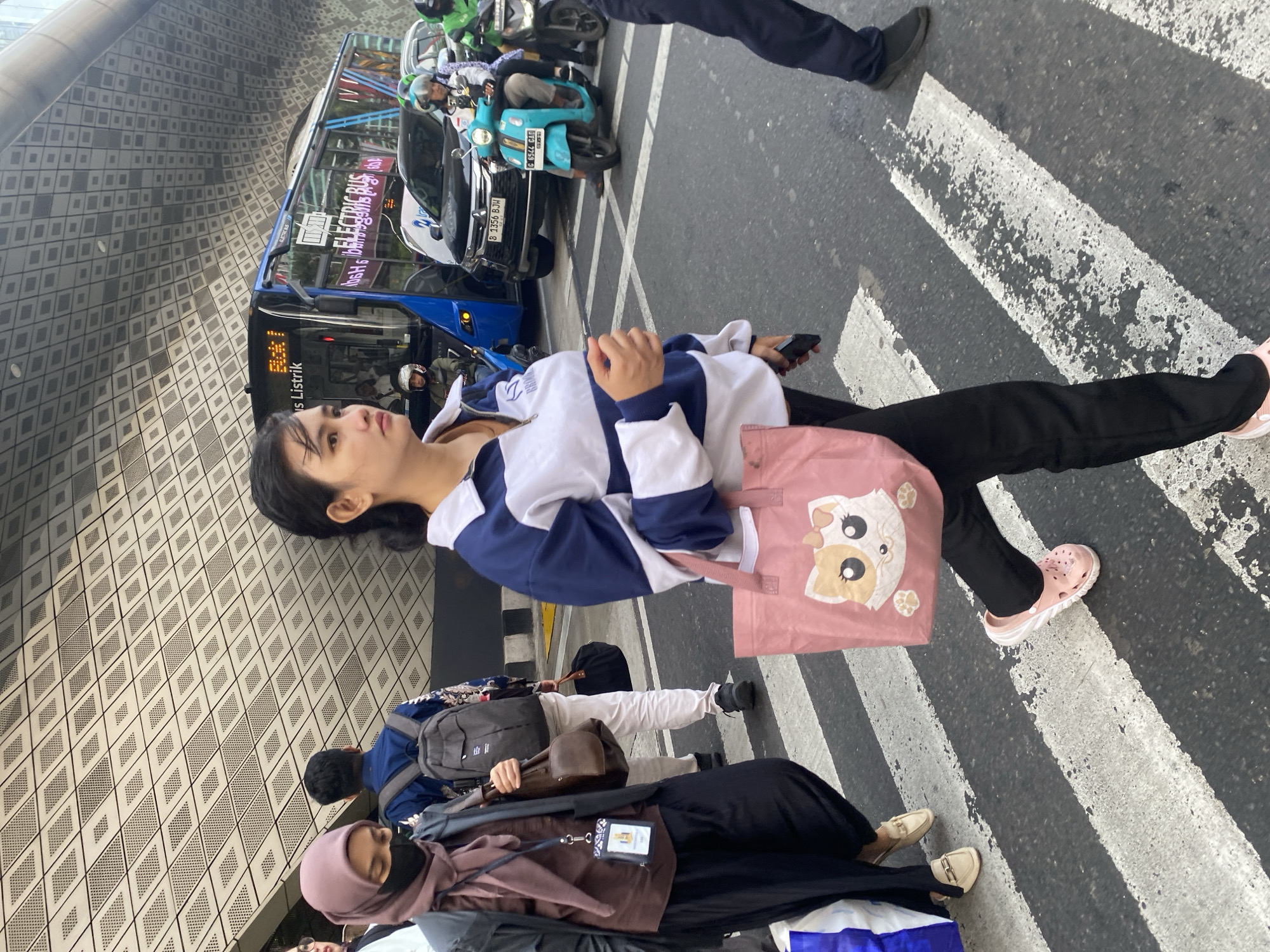 An image taken on a crosswalk in Central Jakarta, Indonesia, shows a woman in the foreground walking right to left, wearing a blue and white striped jacket, black pants, and pink clogs. She carries a pink tote bag with a cat design. Behind her, a woman wearing a pink hijab and black clothing walks left to right. Other pedestrians and various vehicles, including an electric bus, cars, and motorcycles, are visible on the street. The background features a modern building with a patterned facade.