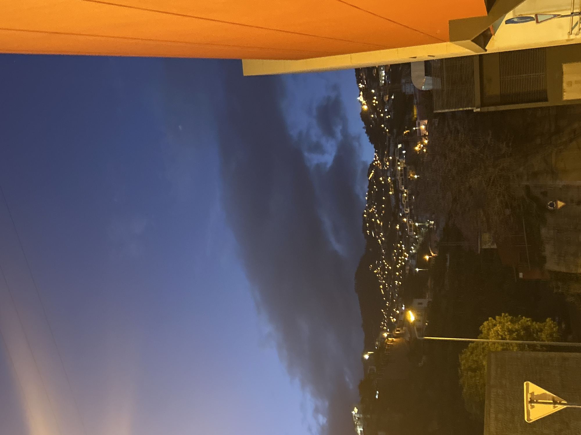The image shows a night view of Câmara de Lobos, Portugal. The sky is dark blue with a large, dark cloud formation. A hillside is visible with numerous small lights scattered across it, likely from buildings or streetlights. In the foreground, there are streetlights, trees, and a road sign with a yellow triangle. The roof of a building is visible at the top of the frame, colored in orange.