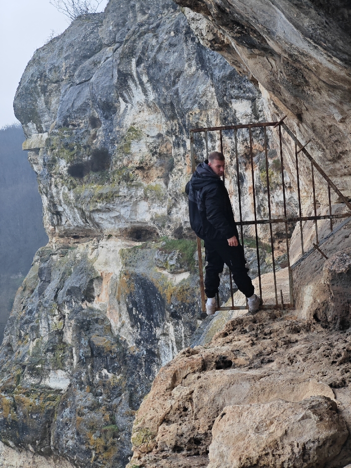 One adult male is depicted standing on an uneven rock ledge within a large, weathered rock formation on the Shumen Plateau, near Shumen, Bulgaria. The individual, appearing to be in his 20s or 30s with short, light-colored hair, is wearing a black hooded puffer jacket, dark trousers, and light-colored athletic shoes with white soles. He is positioned with his back partially turned to the camera, his head rotated to look over his right shoulder directly towards the viewer. His left foot is positioned further back and slightly higher on the rugged rock surface, while his right foot is forward. A rusty, dark-colored metal barrier composed of vertical bars extends from the left mid-ground to the right, angling upwards, separating the subject from the immediate foreground rock. This barrier is secured into the natural rock face. The immediate environment consists of rough, eroded light-brown rock under a natural overhang. To the left and in the background, a massive, multi-toned grey and white cliff face, marked with patches of lichen and sparse vegetation, drops sharply. A hazy, forested valley is visible in the distant background on the far left under an overcast sky.