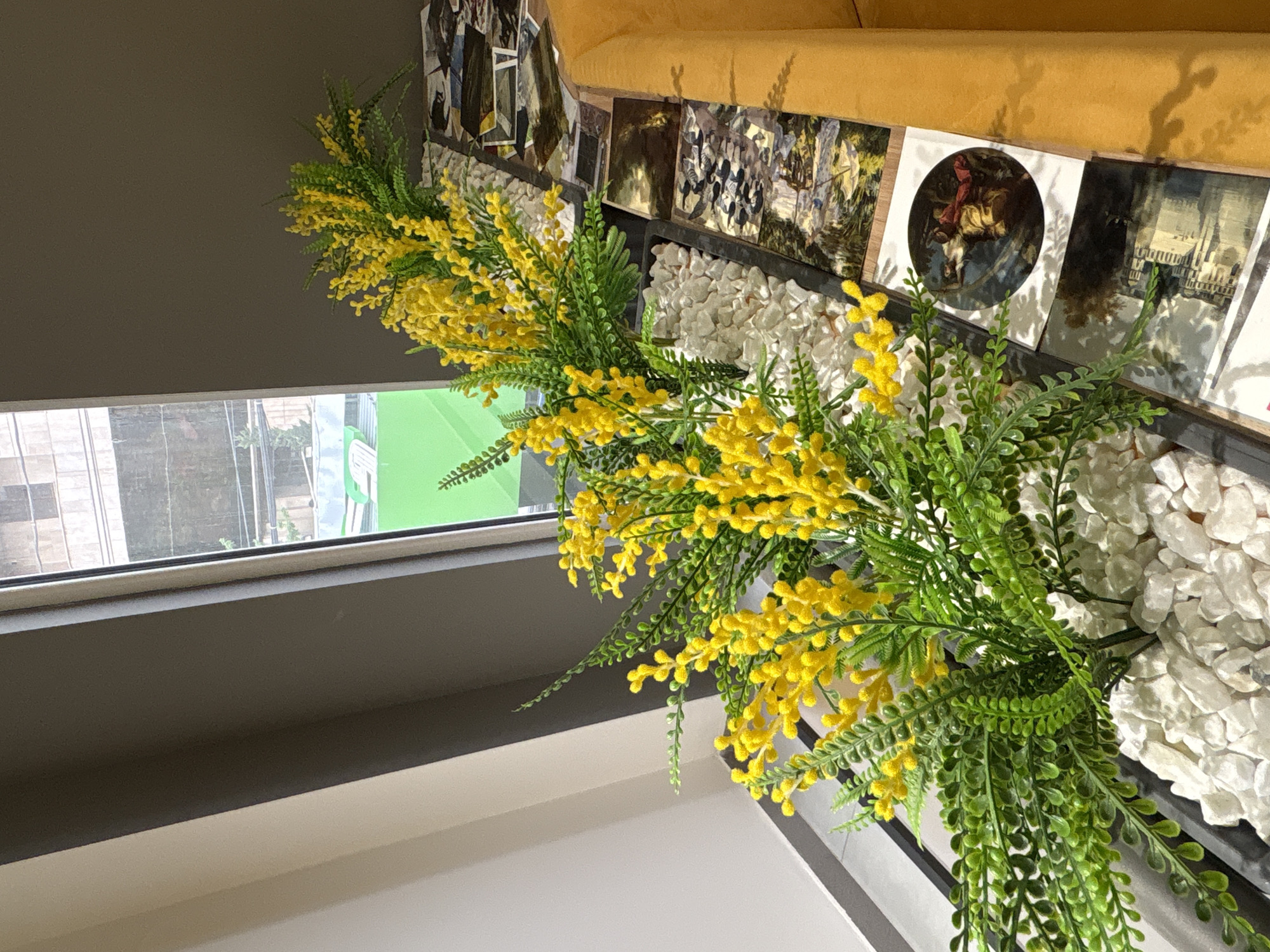 The image shows an indoor setting with a window, a floral arrangement, and decorative elements. The window offers a view of buildings, suggesting an urban environment. The floral arrangement features yellow flowers with green foliage. It is placed on a surface covered with white stones, alongside decorative cards and a yellow sofa. The location is not discernible from the image alone. Therefore, the reference to Yerevan, Armenia, cannot be confirmed based on the provided visual information.