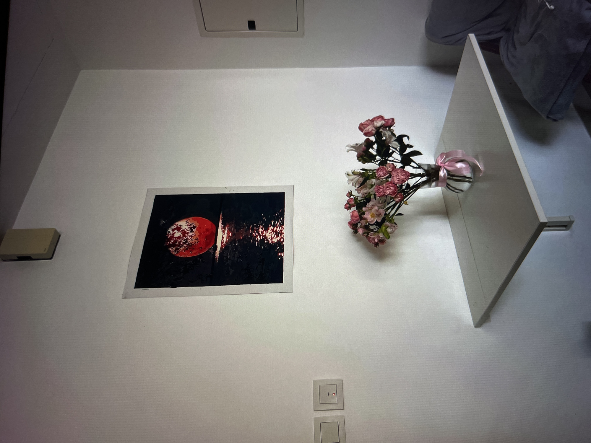 A framed painting of a red full moon reflected on water hangs on a white wall. Below the painting, a vase of pink and white flowers with a pink ribbon sits on a white folding table. To the left of the table are two light switches, one with a red indicator light. To the right of the table, a grey upholstered chair is partially visible. The scene is indoors, likely a residential or hotel room, and does not provide any specific indicators of its location in Dubai, United Arab Emirates.