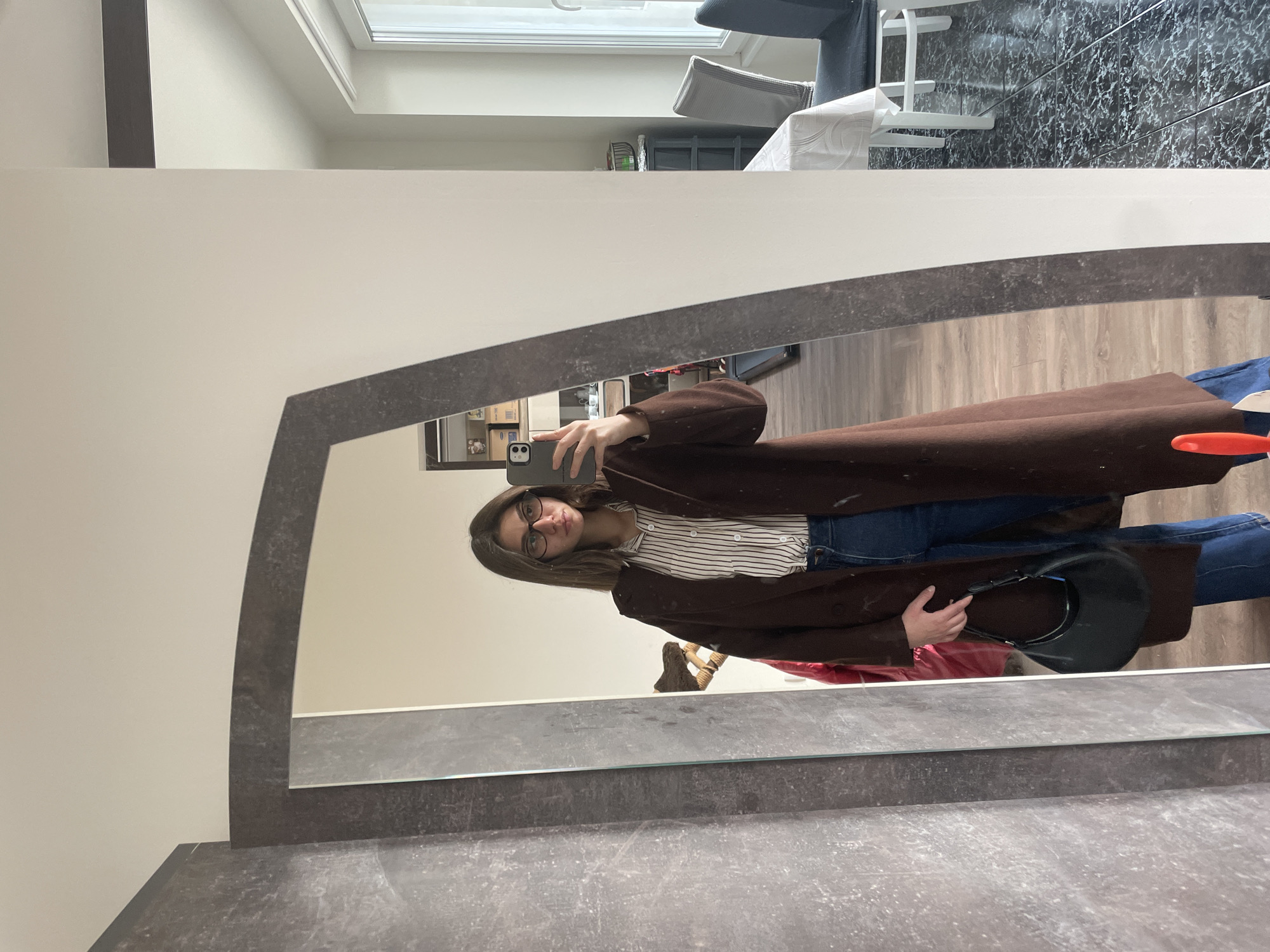 A person stands in front of a large, irregularly shaped mirror, taking a photo with a smartphone. The person is wearing glasses, a striped shirt, blue jeans, and a long brown coat. They hold a black handbag. The background visible in the mirror reflects a room interior. The location appears to be in Sofia, Bulgaria, based on visual architectural cues and interior design elements. The area outside the mirror appears to be a waiting room area in a salon with black, patterned flooring. The image is rotated.
