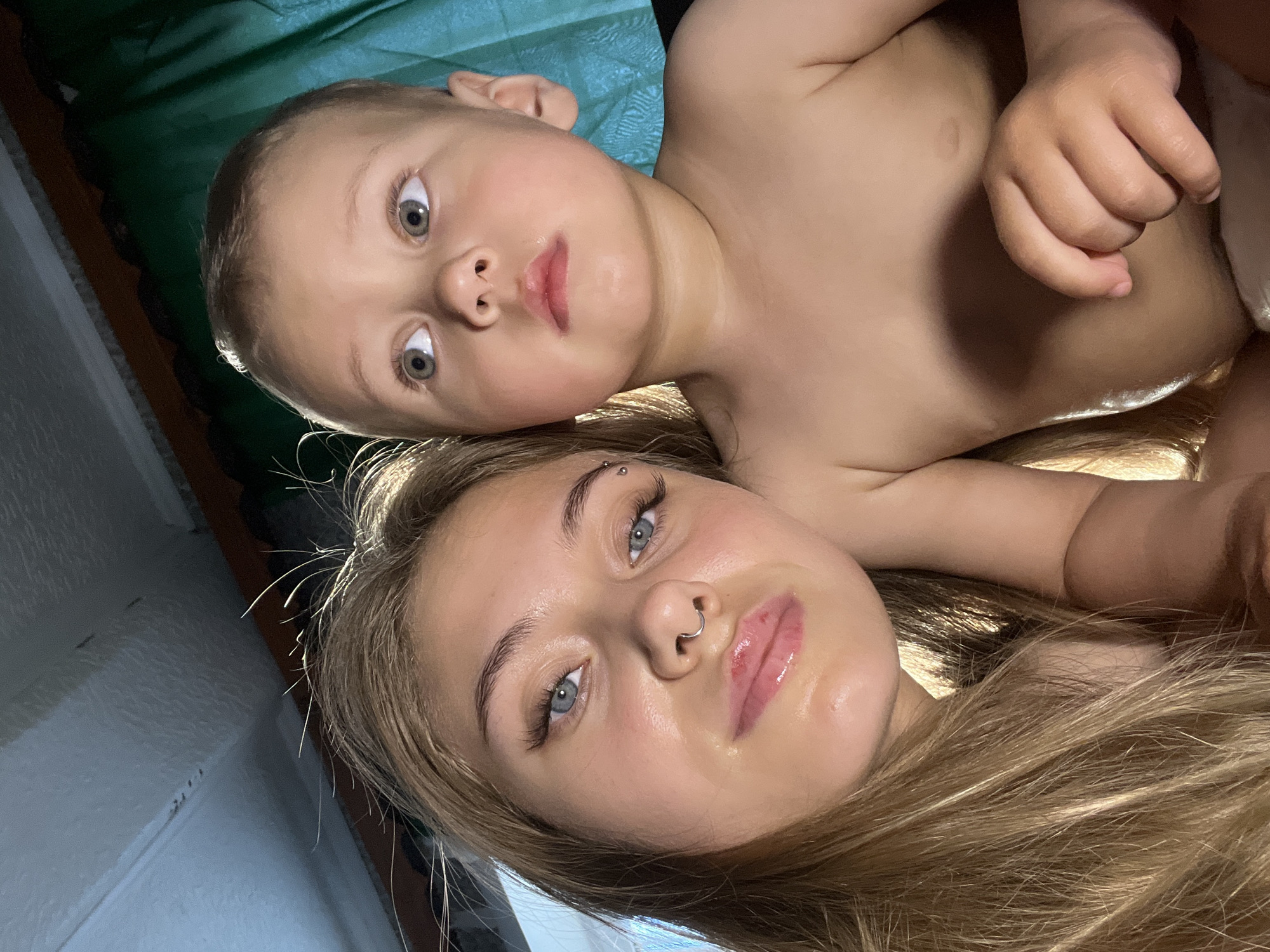 Two individuals, a child and an adult female, are positioned closely together. The child, presumably a toddler, is shirtless and lies above the adult. The female has fair hair and visible facial piercings. They are indoors. Location details about the city of Фоки, Russia, are not evident from the scene.