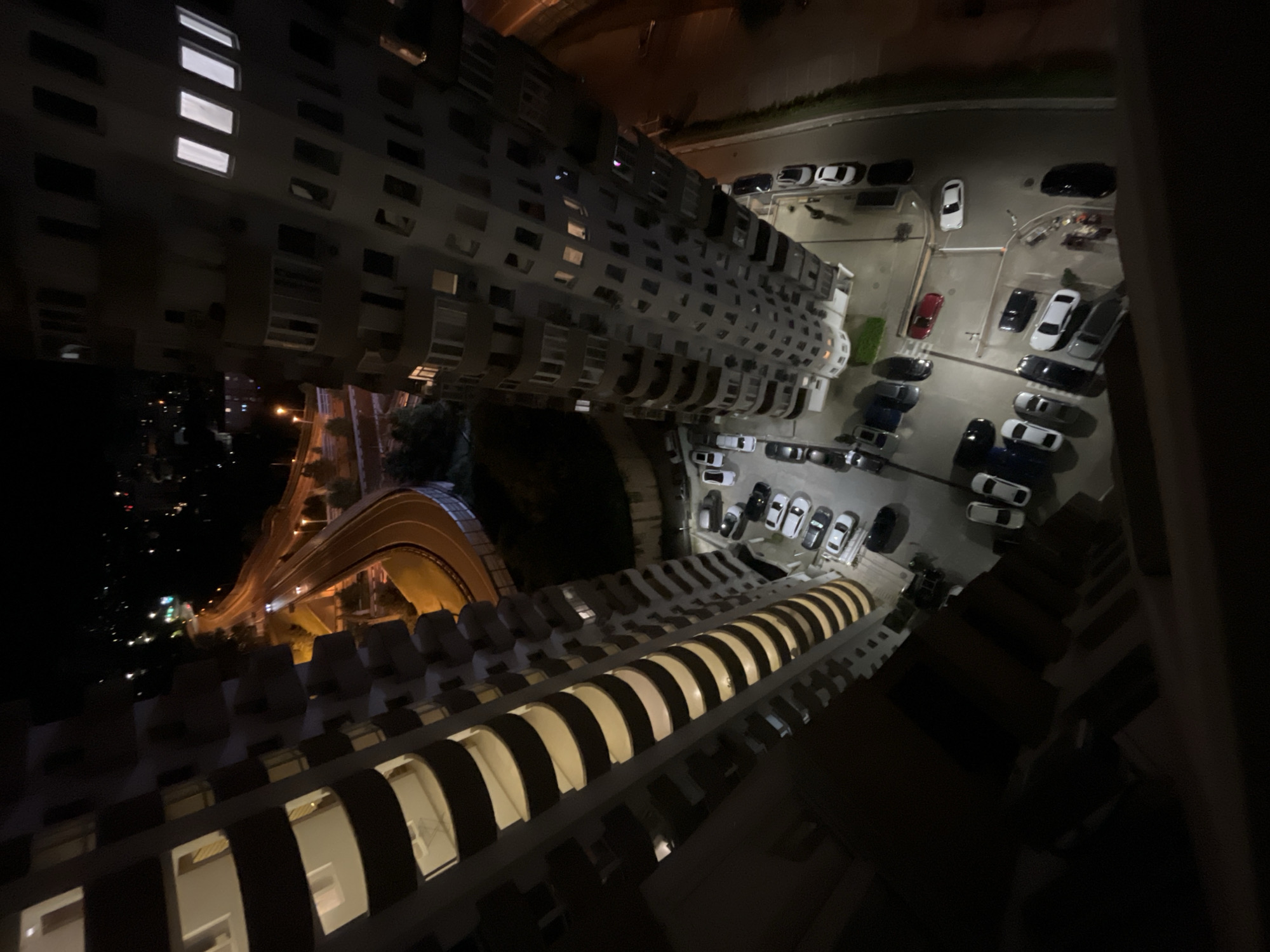 This image presents a high-angle, downward-looking view of an urban residential area at night in Sochi, Russia. The scene is dominated by two tall, multi-story apartment buildings framing the perspective. The building on the left exhibits a facade characterized by numerous curved balconies, some of which are illuminated, revealing interior apartment spaces. The building on the right is also a high-rise structure with multiple windows, several displaying interior lighting.
Between and around these structures are asphalt parking lots, densely occupied by various parked vehicles, including numerous white, black, and silver cars, along with one distinct red car. In one of the parking areas, multiple individuals are discernible: at least one person is seen pushing a stroller or cart, and other figures are standing or walking near parked vehicles, indicating human presence and activity at ground level.
In the mid-ground, a winding, multi-lane road or elevated highway is prominent, illuminated by numerous warm streetlights. The headlights of a single moving vehicle are visible on this road segment. The background consists of dark areas suggesting natural terrain or less developed zones, interspersed with distant ambient city lights. The overall environment is an active, modern urban setting under nocturnal conditions.