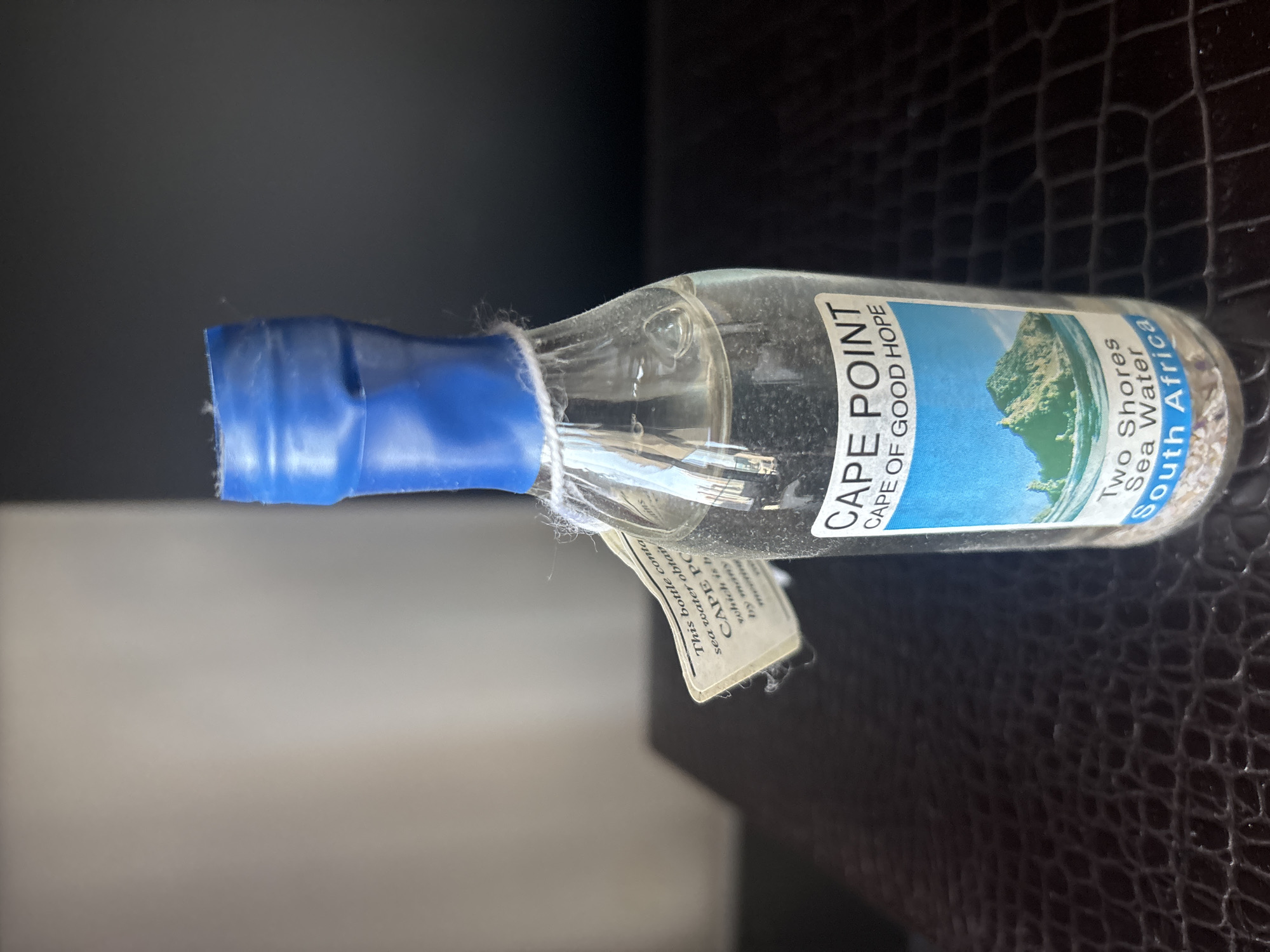 A souvenir bottle containing sea water from Cape Point, South Africa, is centrally framed against a dark, textured surface. The bottle has a blue plastic cap secured with white string. A small tag attached to the neck of the bottle reads "This bottle contains sea water obtained at CAPE POINT, which is h... by many...". A label on the bottle itself displays an image of a mountainous coastline with the text "CAPE POINT", "CAPE OF GOOD HOPE", "Two Shores Sea Water", and "South Africa". The background consists of a split color gradient, with a light grey on the left and a dark grey/black on the right. The scene is lit from the front, casting subtle shadows. There are no people or animals present.