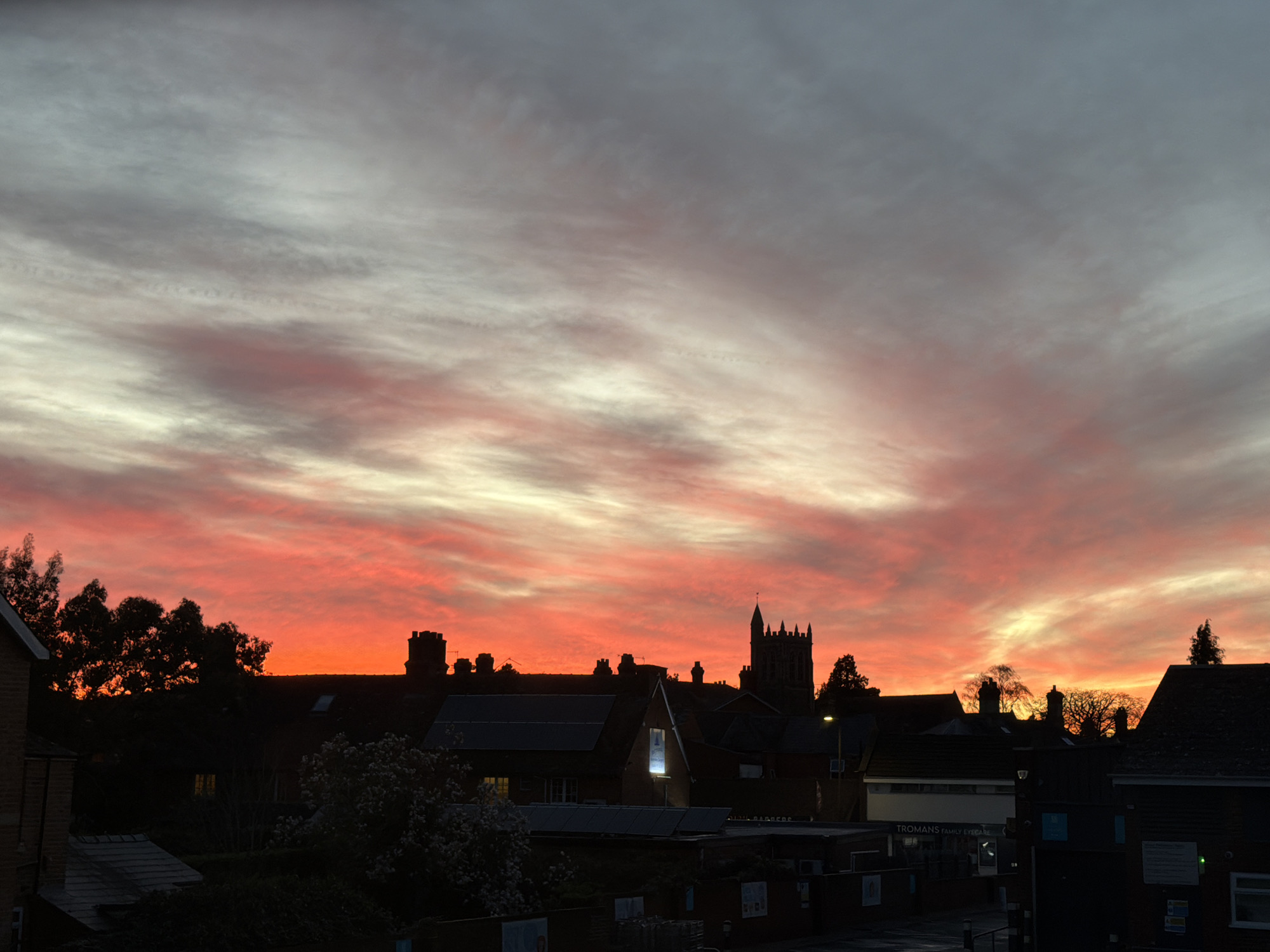 The image captures a sunrise or sunset over a townscape, likely taken in Malvern Hills, United Kingdom. The sky is a vibrant mix of red, orange, and gray hues, indicative of the sun's position near the horizon. Silhouetted against this colorful backdrop are rooftops, trees, and a church tower, revealing a typical European urban setting. The scene is static, lacking any evident human presence or movement. The lighting suggests a time near dawn or dusk, and the overall environment conveys a sense of urban serenity amidst natural beauty.