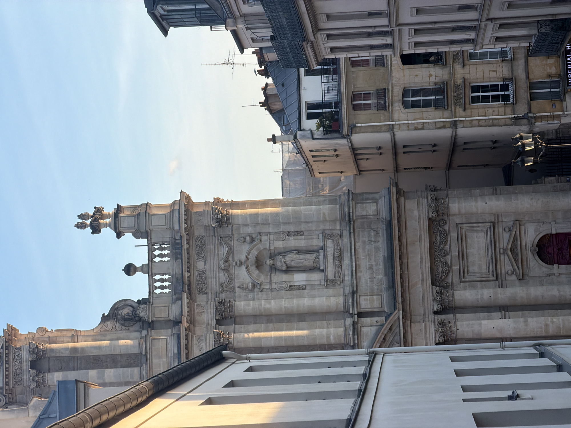 The image shows a section of the Église Saint-Roch in Paris, France, viewed from a low angle. The church's facade is made of light-colored stone and is adorned with intricate carvings and architectural details. A statue is visible within a niche on the upper part of the facade. Adjacent buildings of various heights and architectural styles flank the church, revealing the dense urban environment. The sky is visible above the buildings.