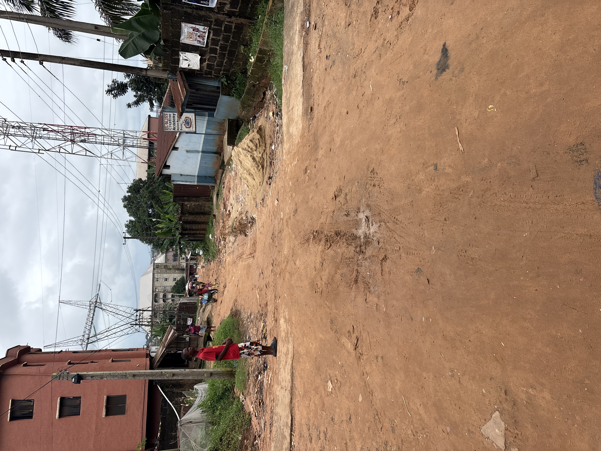 A young boy in a red shirt and patterned shorts stands on a dirt road in Nkpor, Nigeria. He is looking to his left, facing away from the camera. The road is unpaved and appears to be under construction or in disrepair, with tire tracks and puddles of water visible. In the background, there are buildings, including a multi-story red building to the left and a blue and white building with a sign that reads "EL-BLISS Creative Academy" to the right. There are also power lines and a tall transmission tower in the distance. Several people are walking or riding bicycles on the road, and some are carrying goods. A pile of sand is visible on the right side of the road, near the blue and white building. The sky is overcast, suggesting a cloudy day. The scene depicts a typical street scene in an urban area of Nigeria, with a mix of residential and commercial buildings, infrastructure, and daily life.