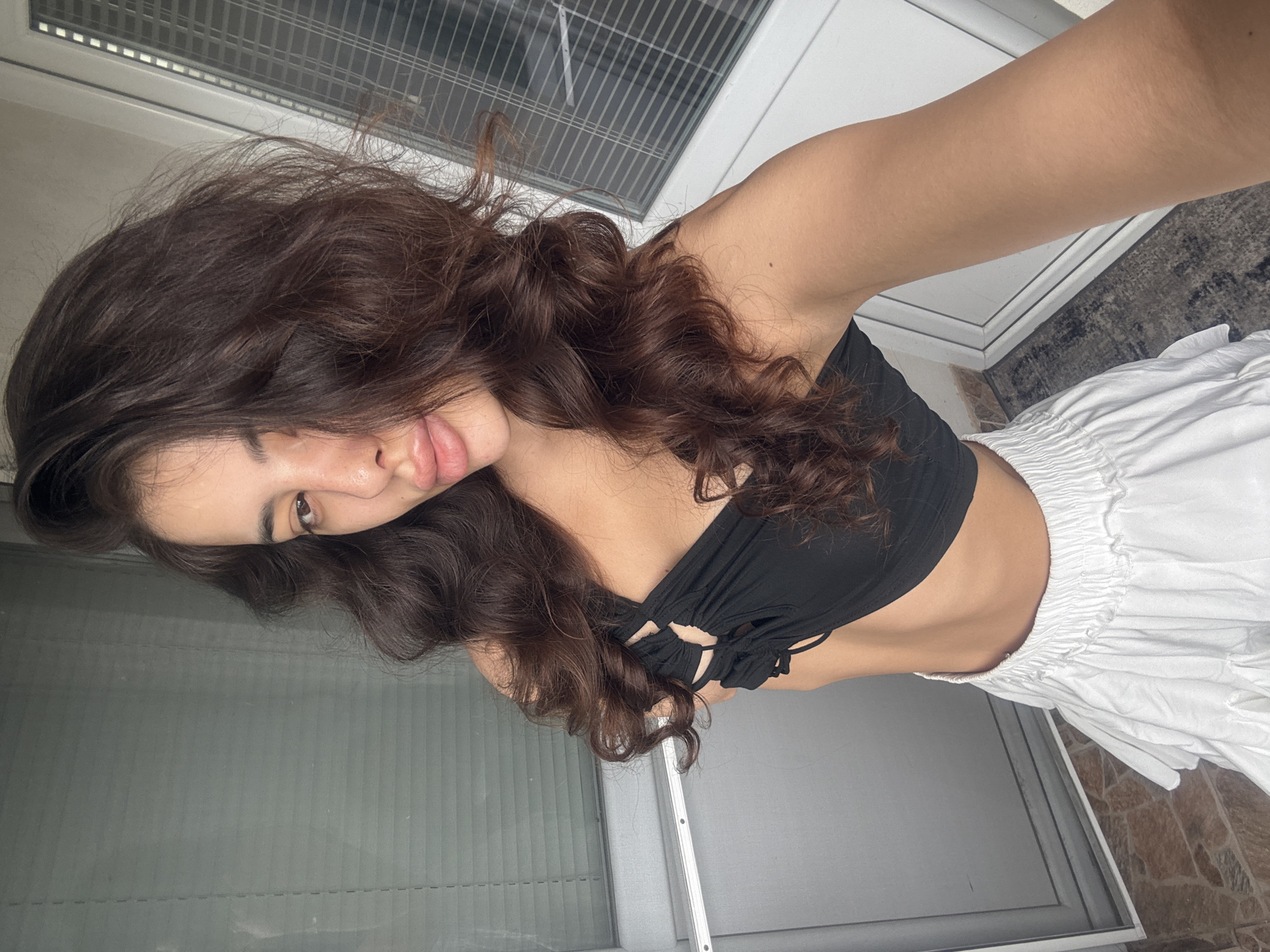 This is a medium shot, selfie-style photo of a young woman with long, dark, wavy hair, taken from a slightly low angle. She is wearing a black, halter-neck bikini top with circular embellishments and white, ruffled shorts. Her left arm is extended towards the camera, and her body is angled towards the right side of the frame. She is looking towards the camera with a slight smile and her lips are parted.
The background appears to be an outdoor setting, possibly a patio or balcony. There is a large window or glass door with blinds or shutters behind her on the left. To the right, there is a glimpse of a floor with a patterned rug and what looks like a tiled area. The lighting suggests it is daytime, with bright sunlight illuminating her skin.
The overall mood of the image is casual and relaxed, with the woman appearing confident and at ease. The image was taken in Plovdiv, Bulgaria. There is no visible text in the image.