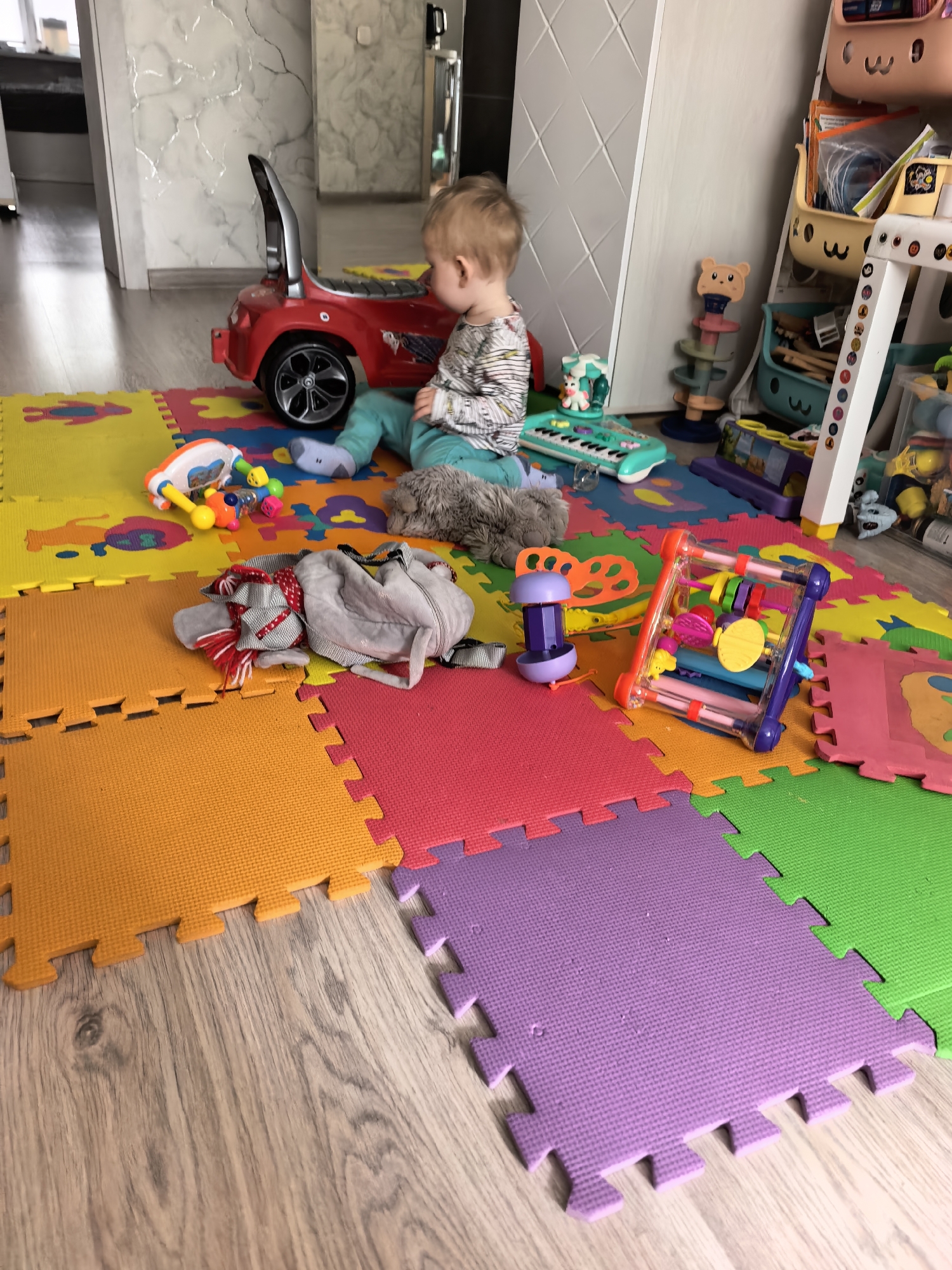 A child sits on a multicolored interlocking foam mat surrounded by toys in an indoor setting. A red toy car is positioned to the left of the child, near a toy keyboard. In the background is a child's storage unit filled with toys. The flooring is wood-look laminate. The location is Sterlitamak, Russia.