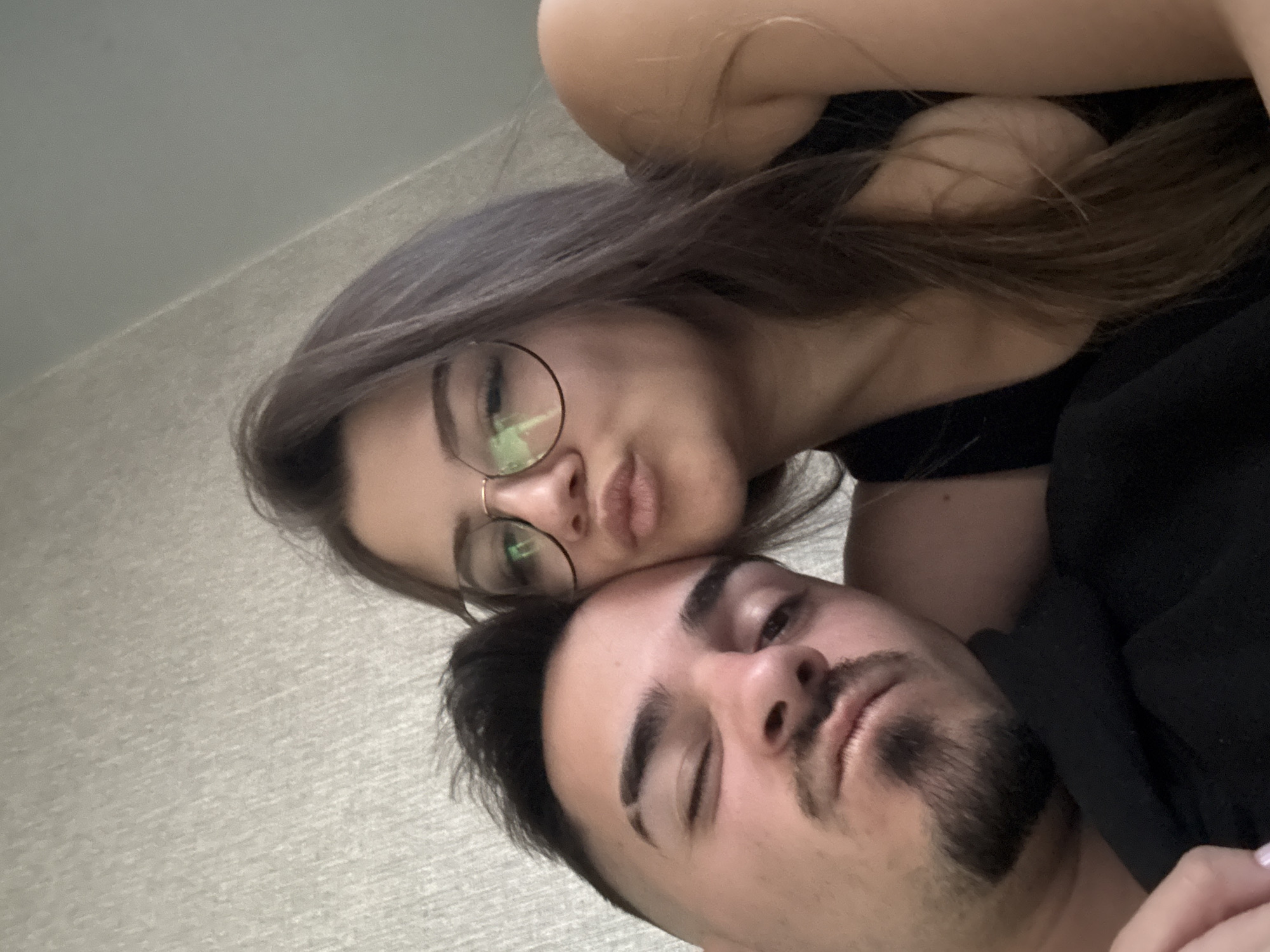 The image depicts a selfie of a woman and a man. The woman, positioned above the man, wears eyeglasses and has long brown hair. She is puckering her lips slightly. The man, lying below her, has dark hair and a beard and is looking up at the camera. Both individuals appear to be wearing dark clothing. The background is a neutral, possibly a wall or ceiling. The location in the city of Sofia, Bulgaria, is not discernible from the image. No city details are visible.