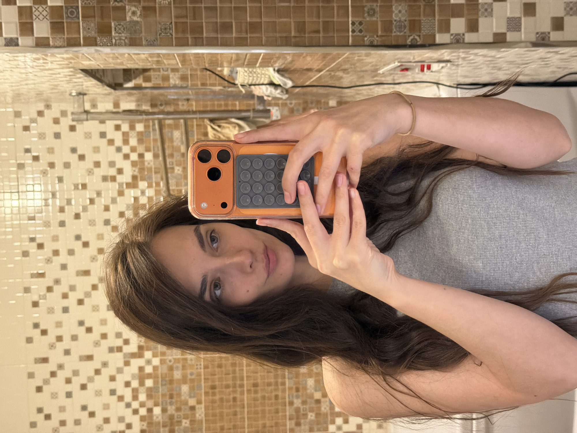 A young woman with long, dark brown hair is taking a selfie in a bathroom. She is wearing a grey tank top. She is holding an orange phone with a black case in her right hand, partially obscuring her face. Her left hand is also visible, holding the phone. The background is a tiled wall with a mosaic pattern of beige, brown, and off-white squares. A mirror reflects part of the bathroom, showing a towel rack and other tiled surfaces. The lighting is bright and appears to be from overhead.
