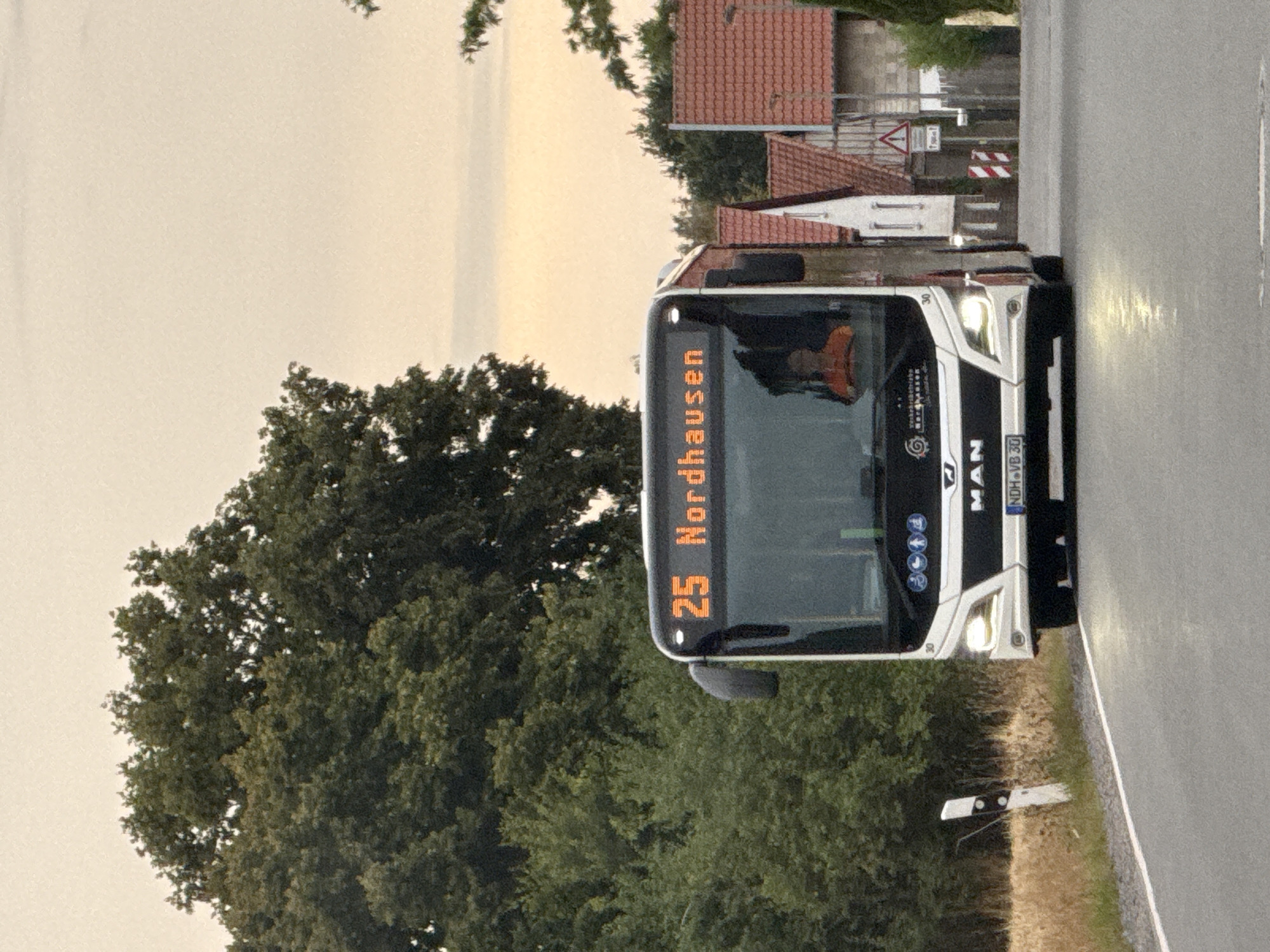 The image shows a white and black MAN bus with an orange digital display showing "25 Nordhausen." The bus is driving on a paved road. The bus has a driver wearing an orange shirt, visible through the windshield. The background includes a line of trees on the left, and a row of houses with red tile roofs on the right. The sky is overcast and pale orange. The scene appears to be in a rural or suburban area. There is a white road marker with black stripes on the left side of the road. The license plate of the bus is visible and reads "NDH-VB 30." There is a traffic sign with a red and white striped pattern and a warning sign on the right side of the road. The image is taken from a frontal perspective, slightly below the eye level of the bus driver.