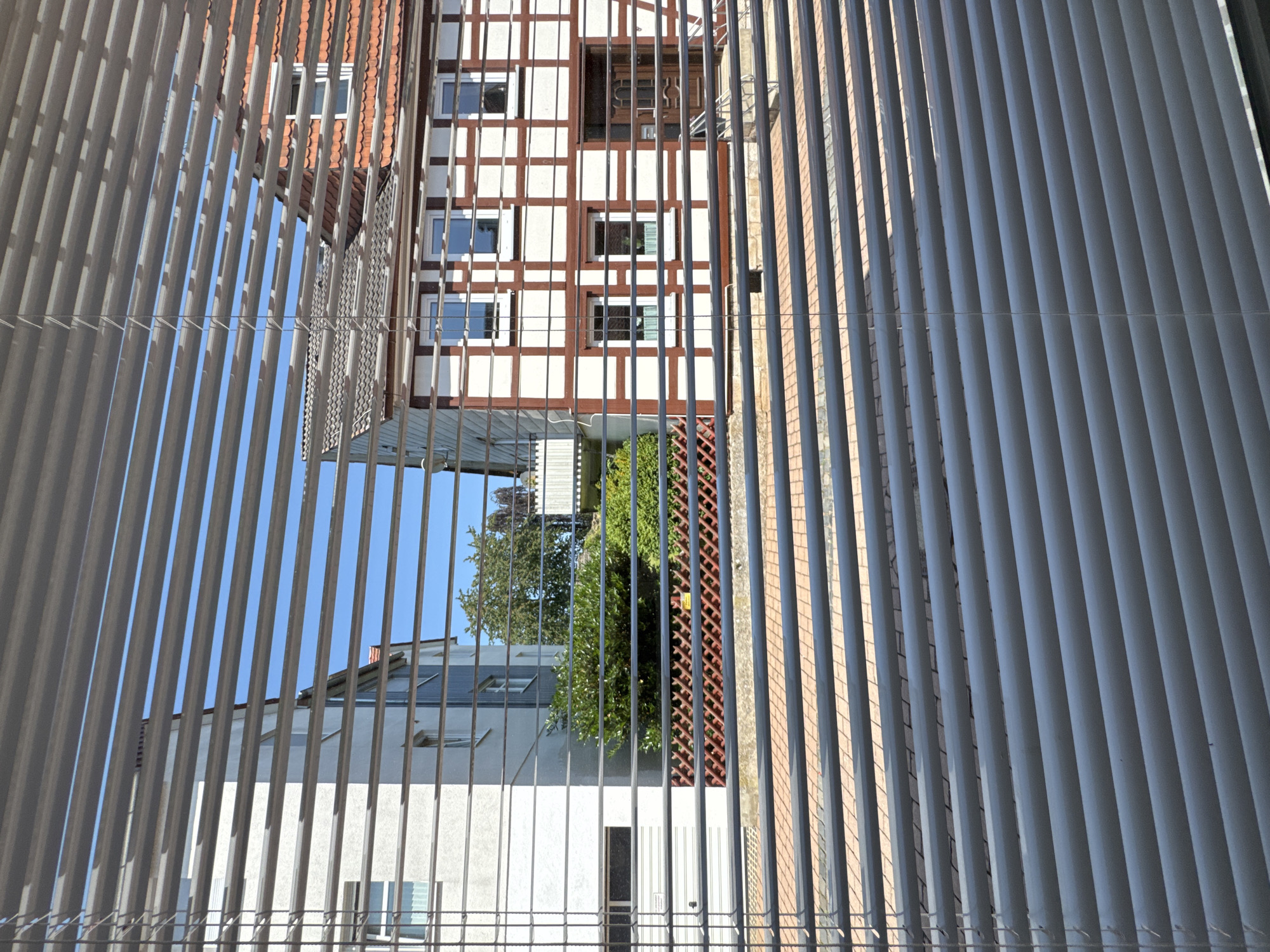The image shows an unusual, inverted perspective of a building exterior, viewed through a set of vertical blinds. The blinds, with their thin, uniform slats, dominate the foreground and create a striped effect that partially obscures the view. The building itself appears to be multi-story and features a traditional timber-framed facade with white infill panels and dark brown structural beams. Several windows with white frames are visible on this section of the building.
Below this, another part of the building is visible, with a lighter colored facade and a sloped roof with visible skylights. A lush green tree is partially visible in the gap between the buildings. The sky is a clear, bright blue, suggesting fair weather. The overall impression is of an architectural study or a unique photographic perspective, possibly looking up from a lower vantage point and inverting the image, or looking down into a courtyard from above. There is no visible text in the image. The scene is static, with no discernible activity. The lighting suggests it is daytime, with clear shadows cast by the blinds and the building.