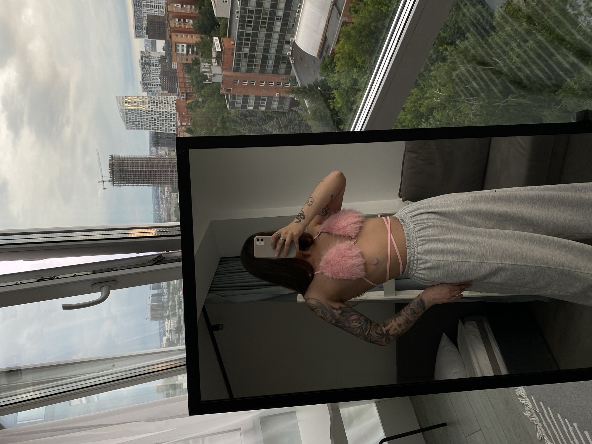 This is a mirror selfie taken by a young woman in what appears to be an apartment. The woman has long, dark hair and is wearing a pink fuzzy bralette with criss-cross straps and gray sweatpants. She has tattoos on her arms and a small crescent moon tattoo on her chest. She is holding a smartphone to take the photo.
The setting is an indoor space with a large mirror reflecting the woman and the room behind her. The mirror also reflects a window with a view of a cityscape. The cityscape shows numerous buildings, some of which appear to be under construction, and lush green trees. The sky is cloudy, suggesting it could be daytime. The overall mood is casual and relaxed.
The view through the window offers location cues of Izhevsk, Russia, with a distinct urban landscape and architectural styles typical of Russian cities. The presence of construction sites indicates ongoing development. The indoor environment seems modern and minimalist, with a grey sofa visible in the reflection.