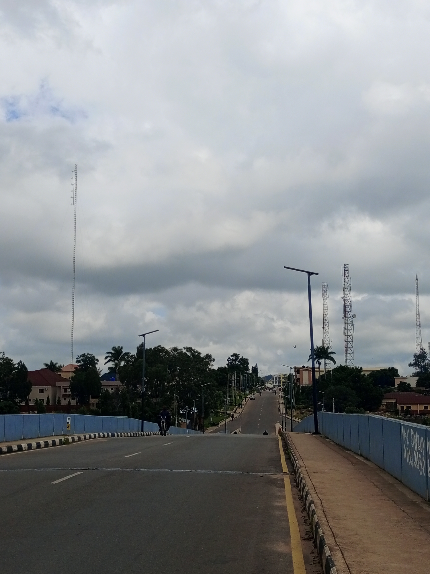 The image shows a view from a bridge overlooking a street in Jos, Nigeria. The bridge has a blue railing with white and black markings along the edge. The road ahead ascends a hill, lined with trees and buildings on either side. Several tall communication towers are visible in the distance. The sky is overcast with gray clouds. A lone motorcyclist is seen riding up the hill on the road. The scene suggests a typical urban landscape in Jos, characterized by its hilly terrain and modern infrastructure.
